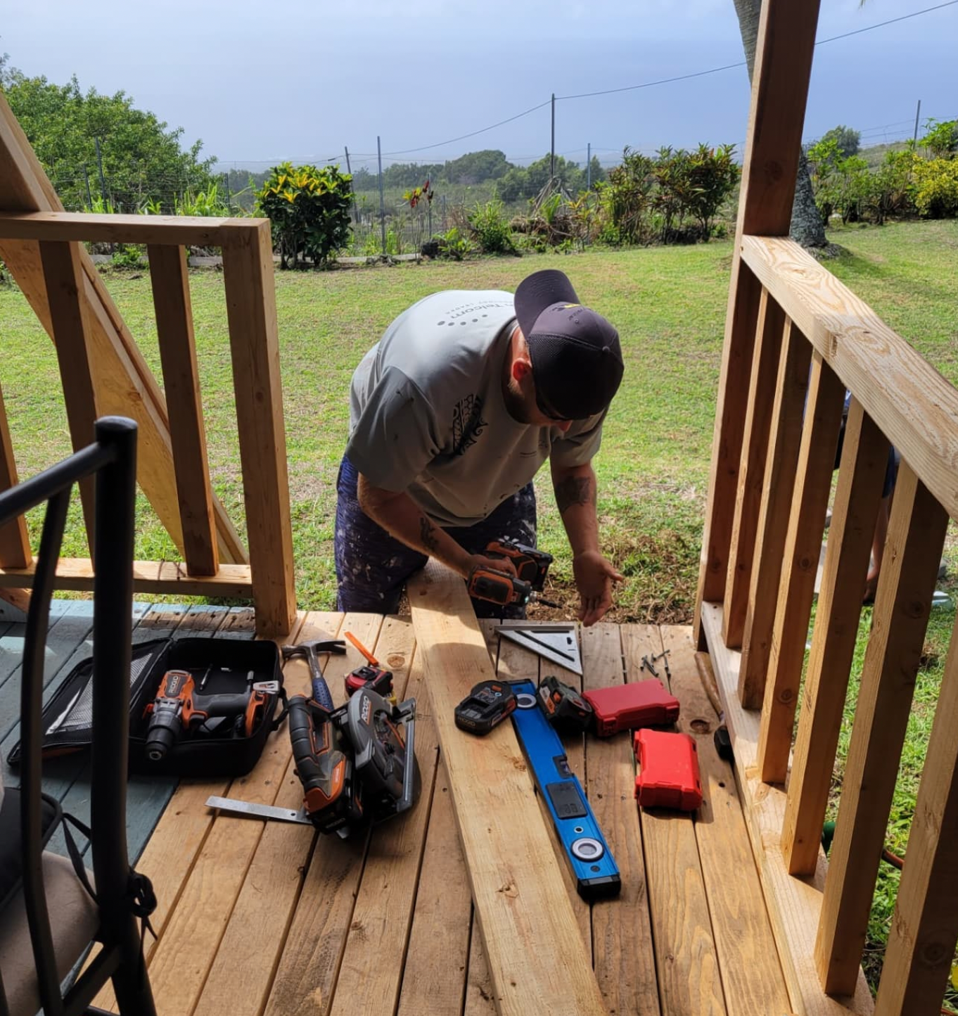 Person kneeling on a wooden porch, using tools beside a blue chainsaw in a sunny yard.