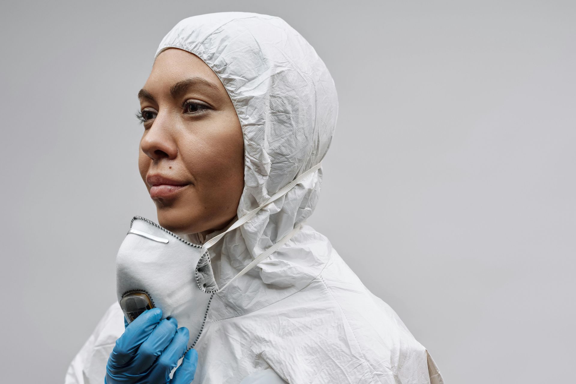 Person in white protective suit and hood, wearing blue gloves, looking to the side. Grey background.