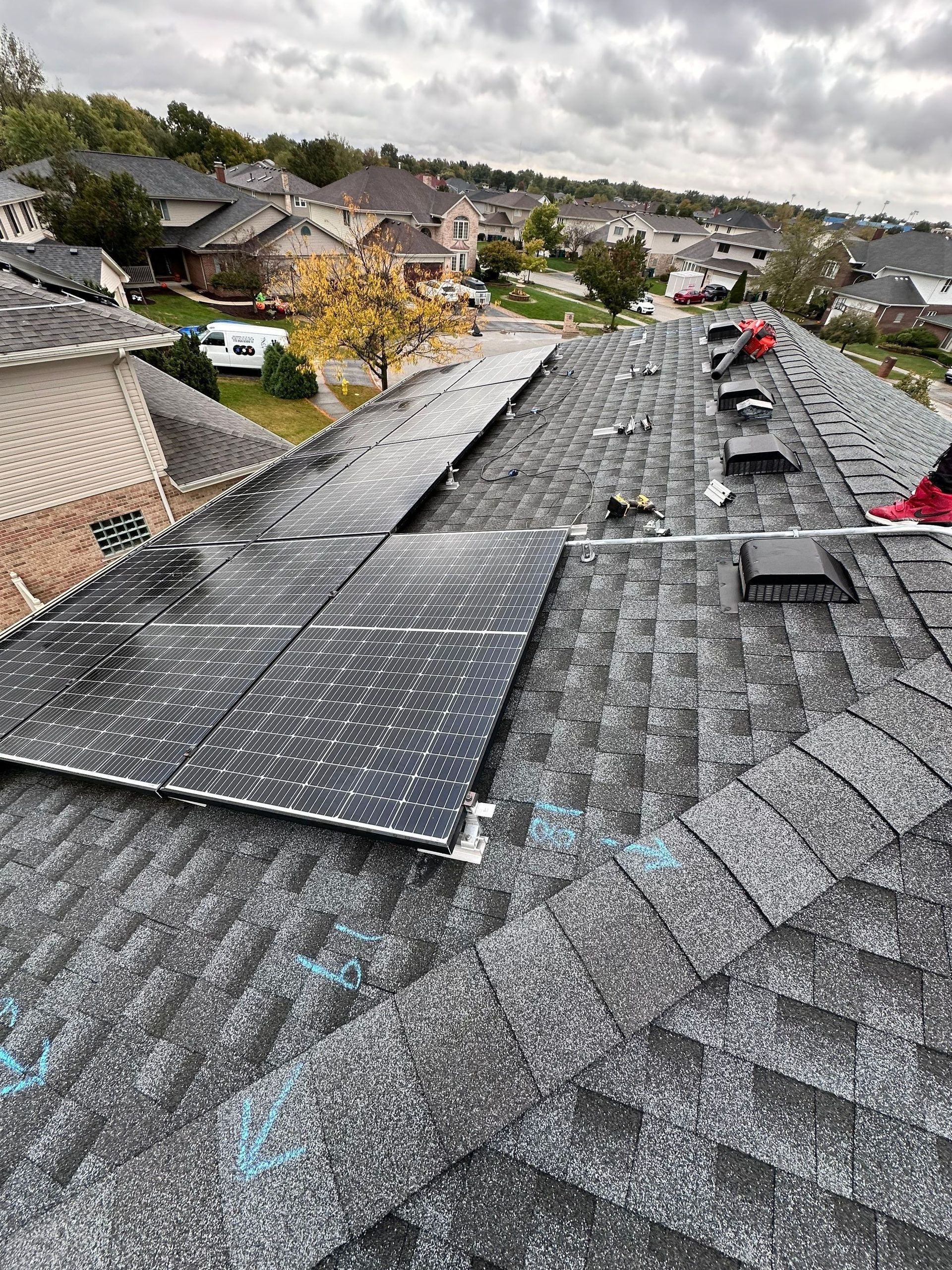 Workers installing solar panels on a shingle roof in a residential neighborhood under a cloudy sky.