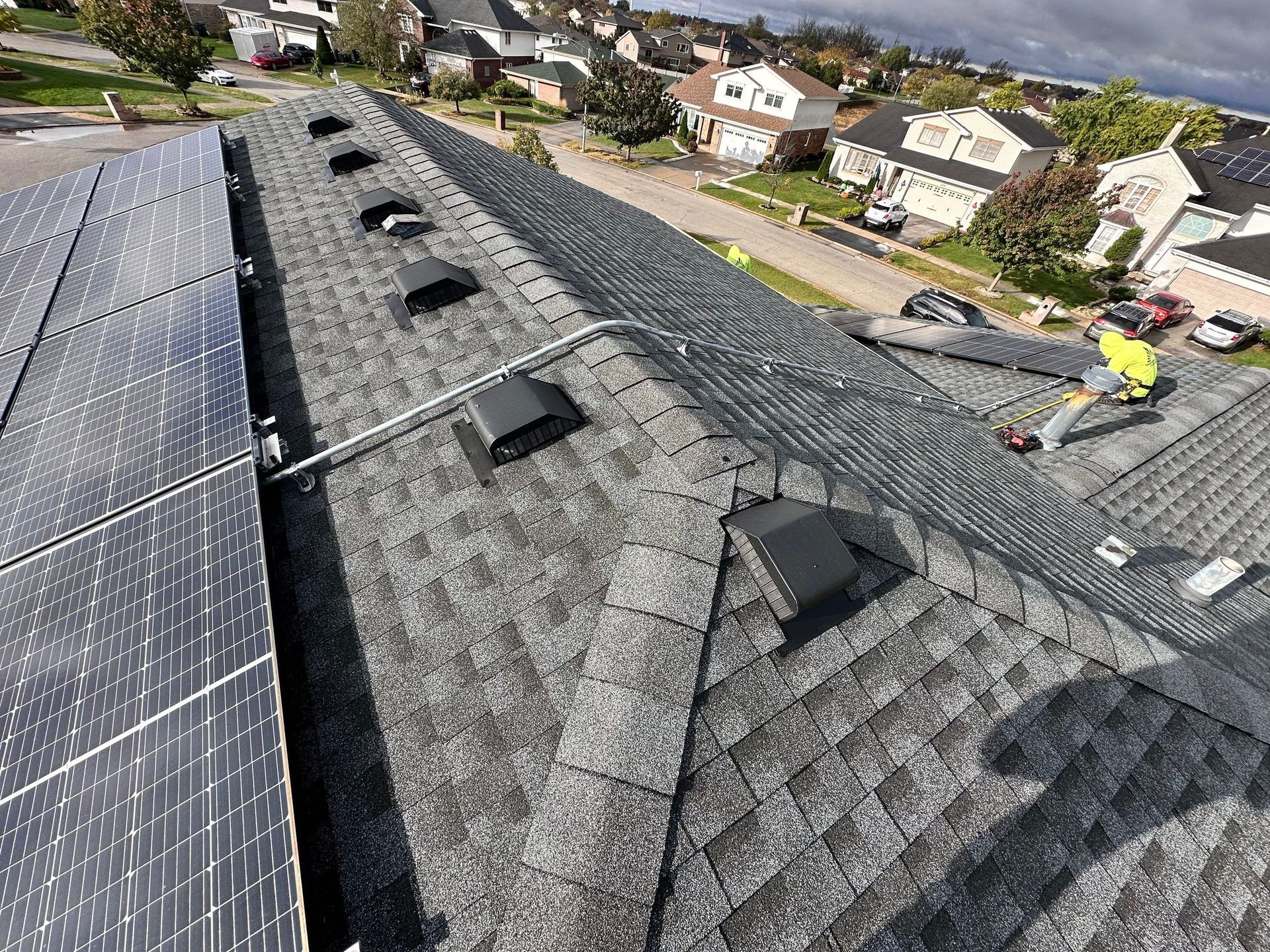 Roofer working on a gray shingled roof with solar panels, vents, and houses in the background.