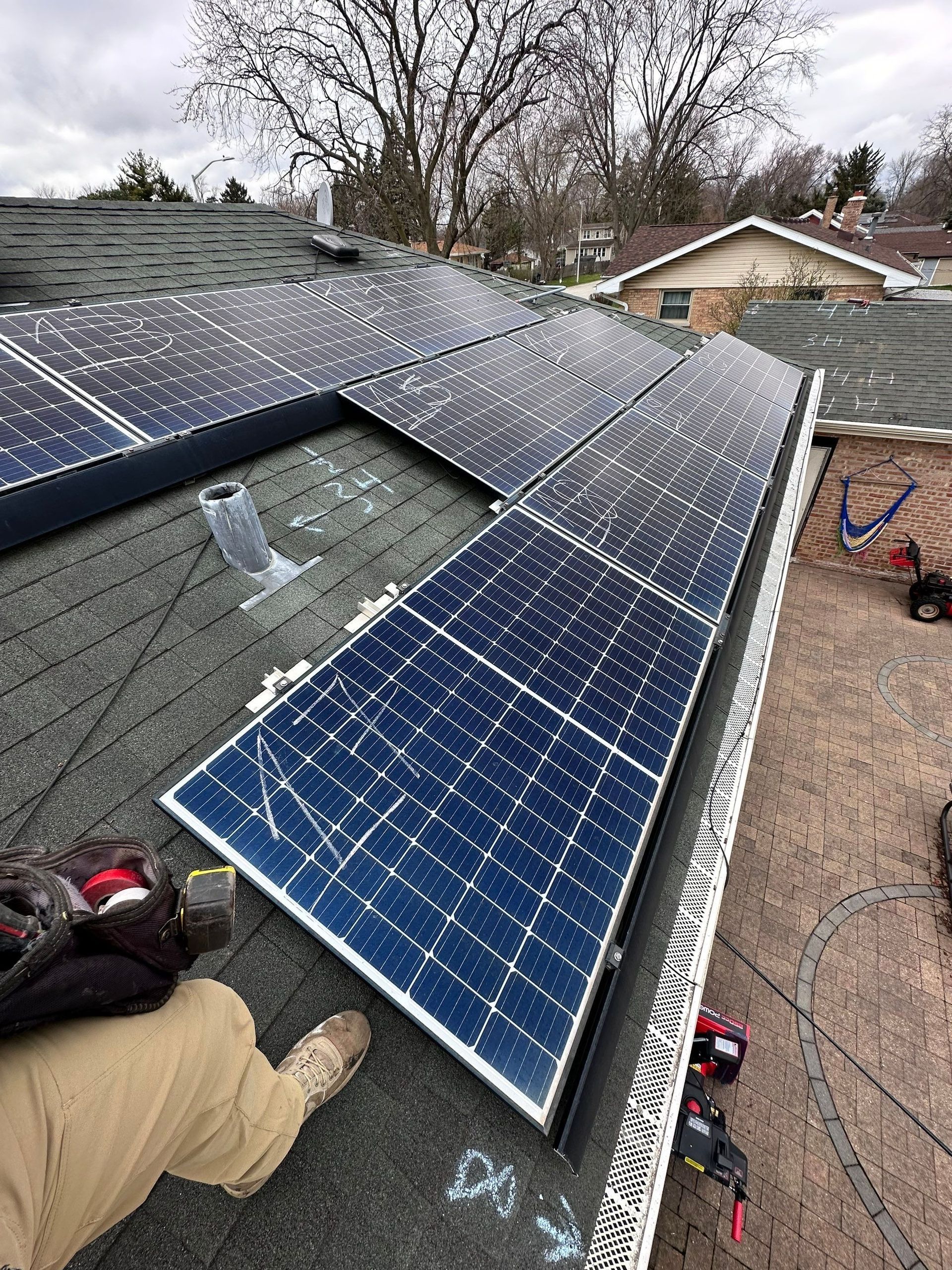 Solar panels installed on a rooftop, partially obscured by a worker in beige pants, with trees and other houses in the background.
