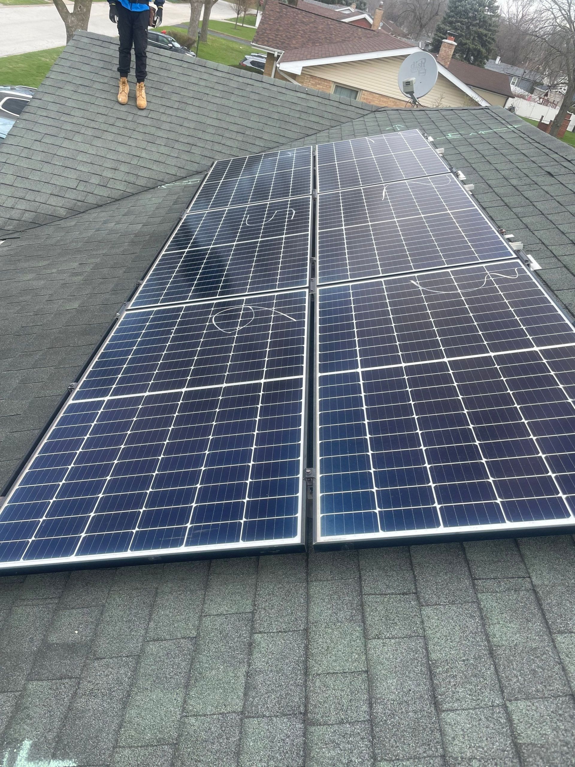 Solar panels installed on a dark shingled roof; person stands nearby.