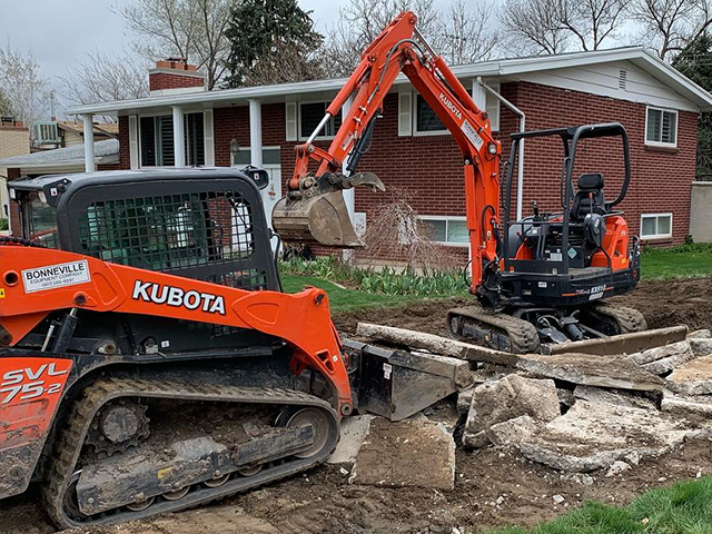 A bulldozer is digging a hole in front of a brick house.
