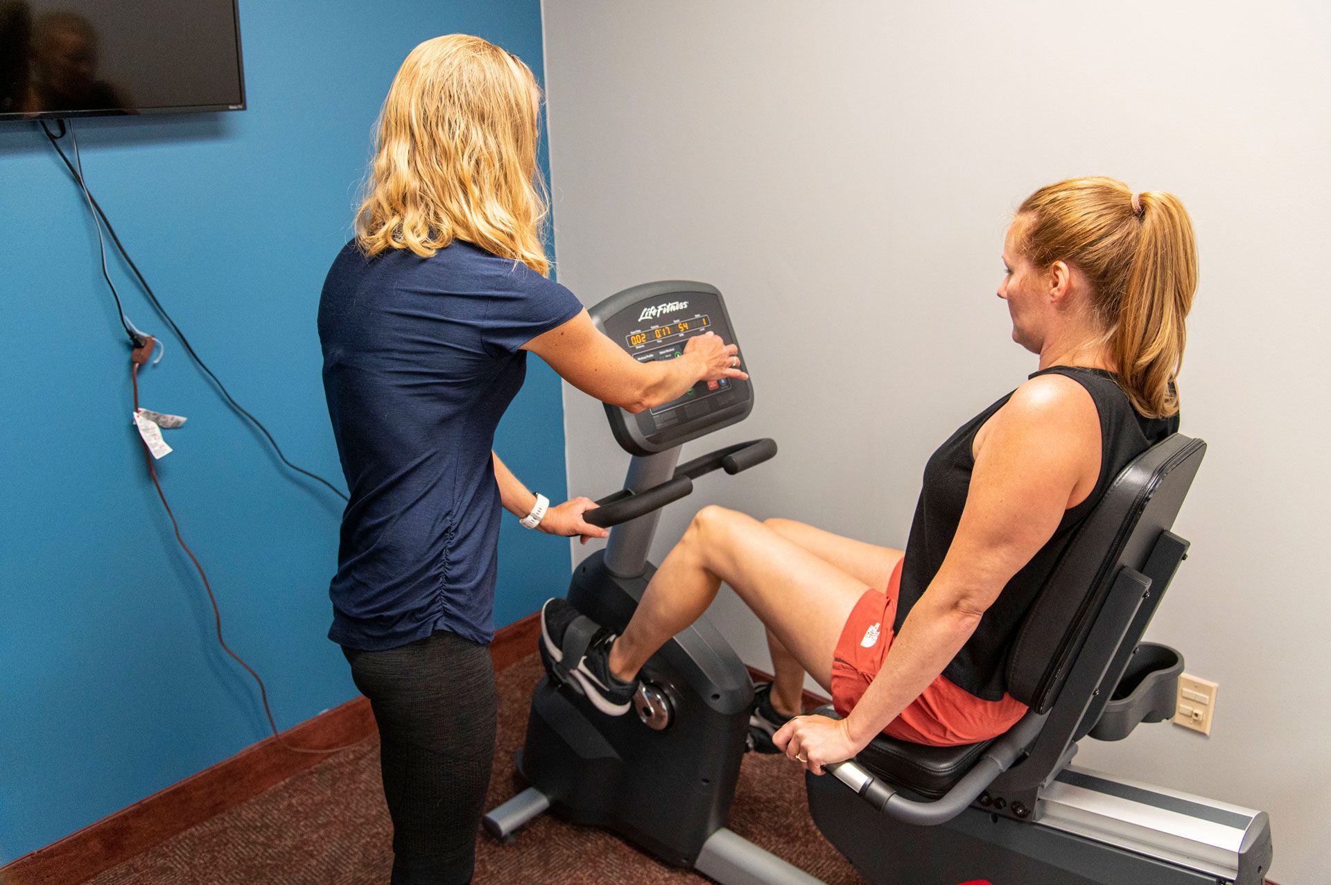 Woman on recumbent bike, guided by another, indoors. Adjusting settings, exercise in progress.