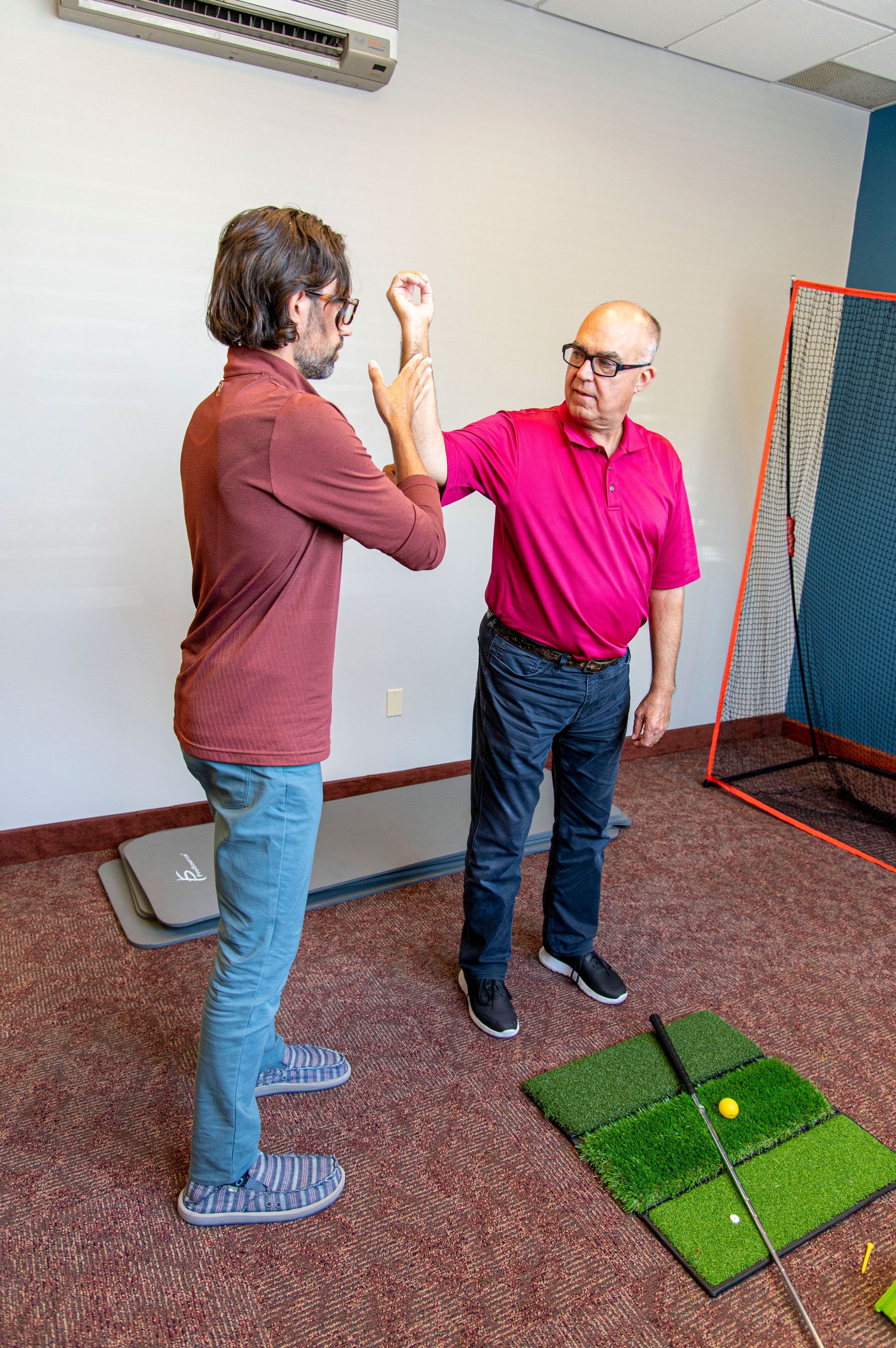 Two men in an office setting. One in red shirt examines the arm of another in maroon shirt and jeans.