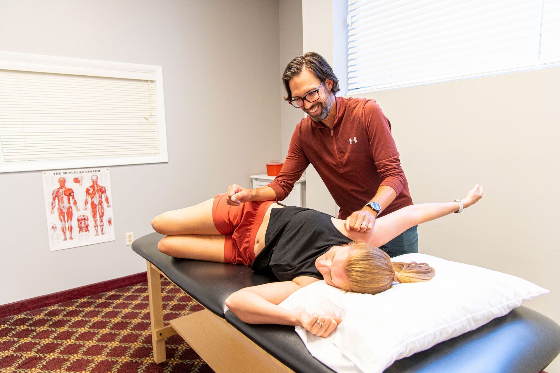 Therapist assisting a patient with arm movement on an examination table. Interior setting with chart on wall.