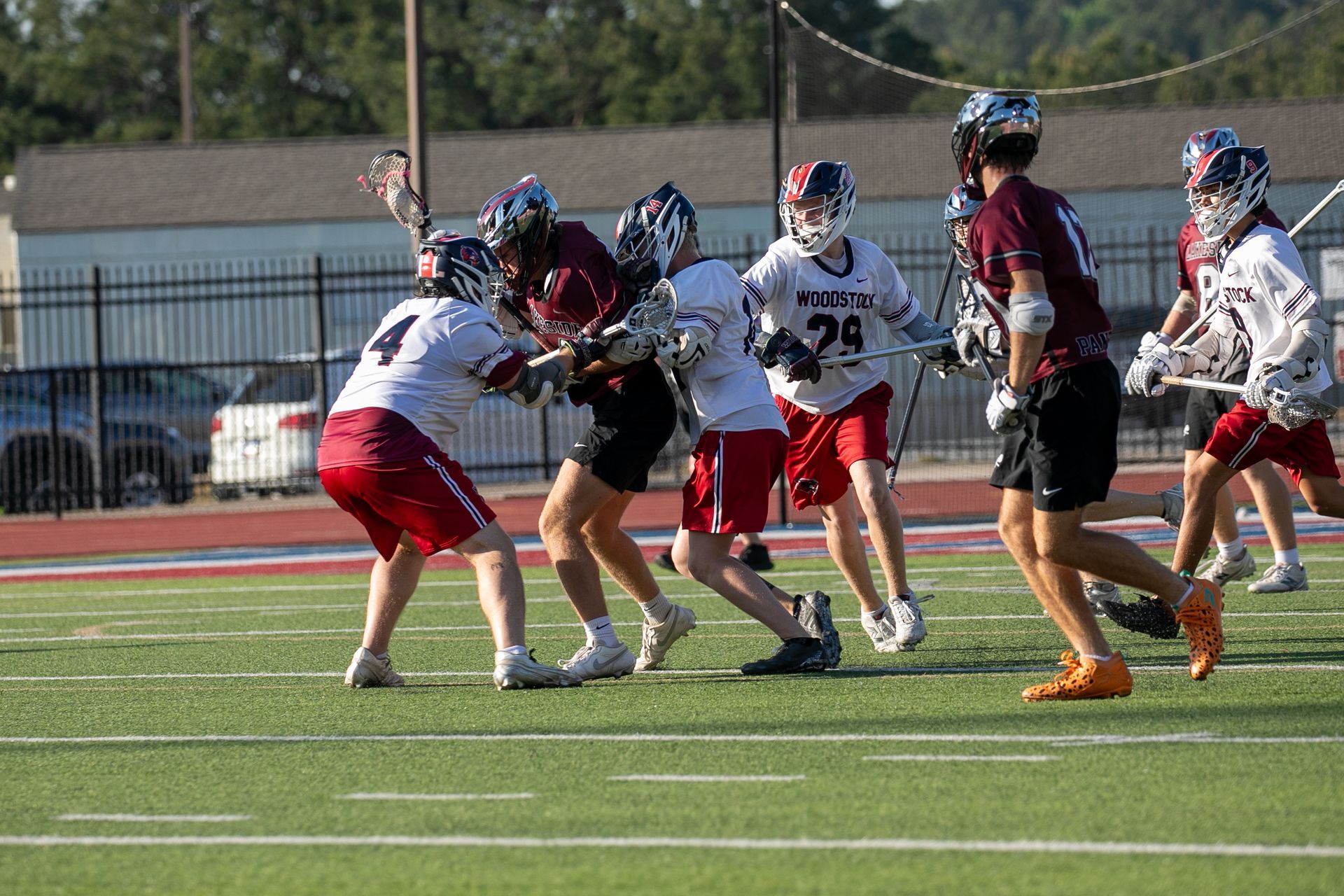 Lacrosse players in white and maroon uniforms battling for the ball on a sunny outdoor field.