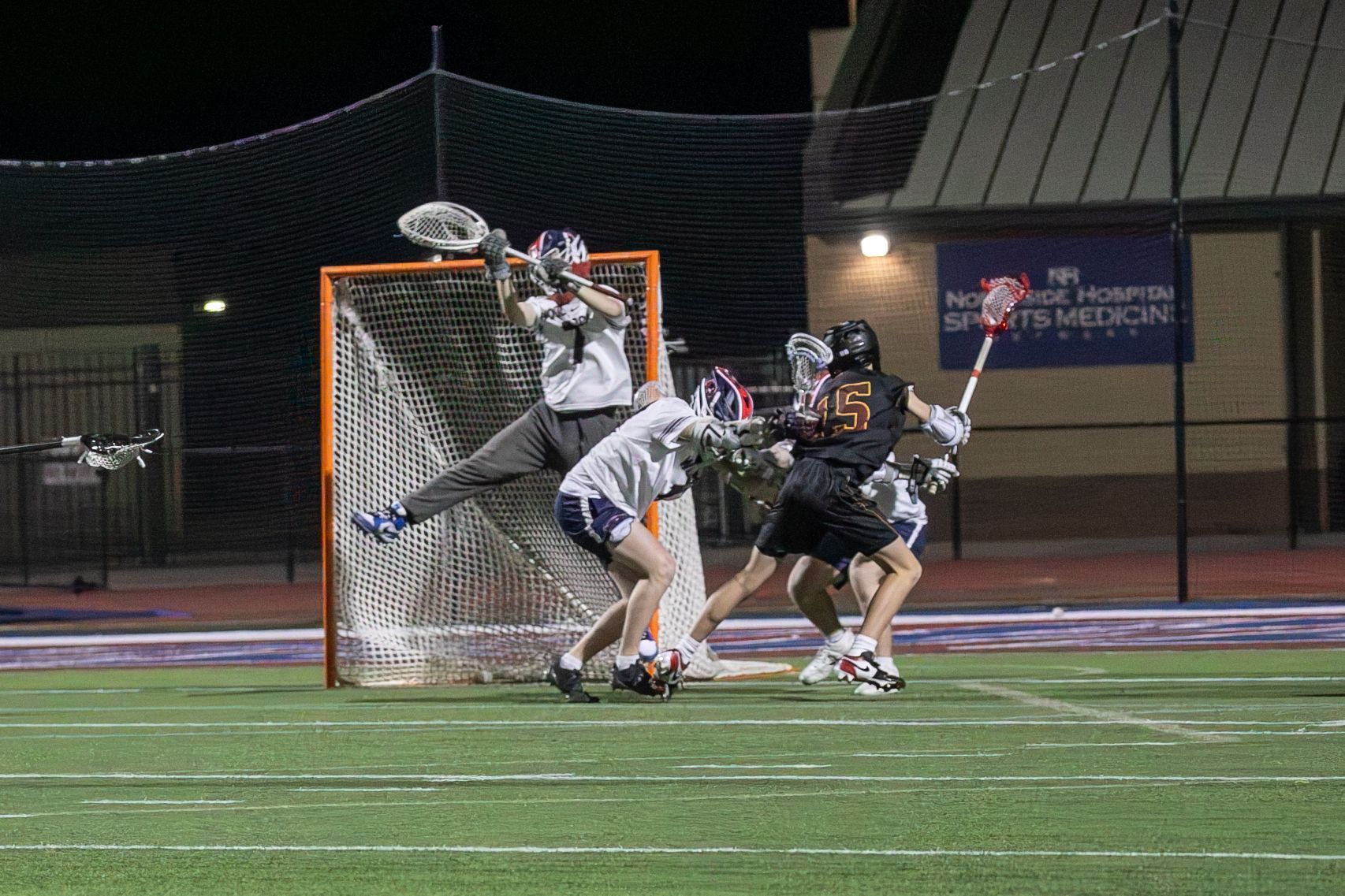 Lacrosse players battling for the ball in front of the goal at night on a lit field.