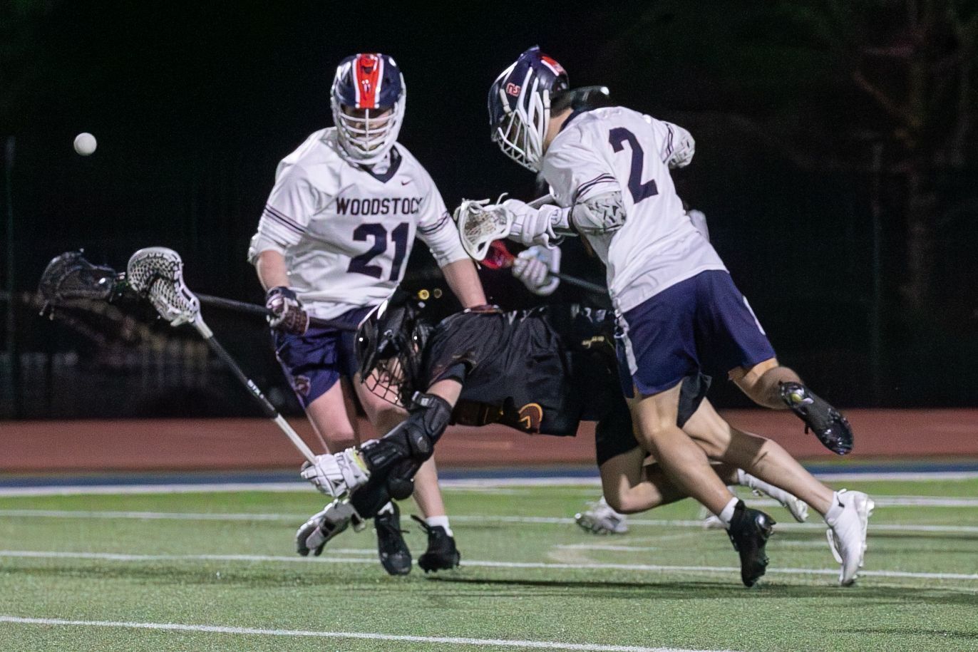 Two lacrosse players in white and dark uniforms chase the ball on a lit field at night