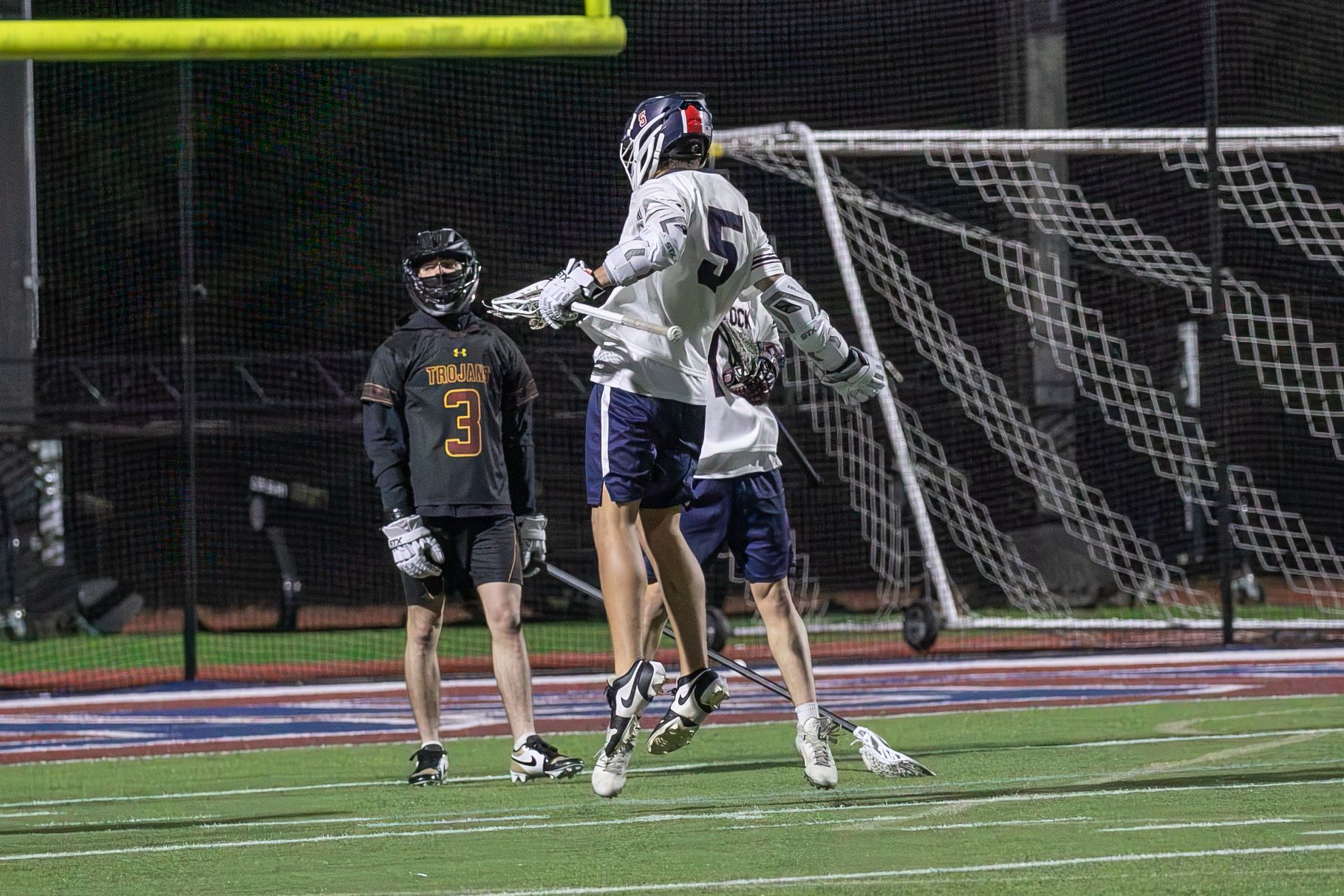 Two lacrosse players jump near the goal during a nighttime game on a turf field.