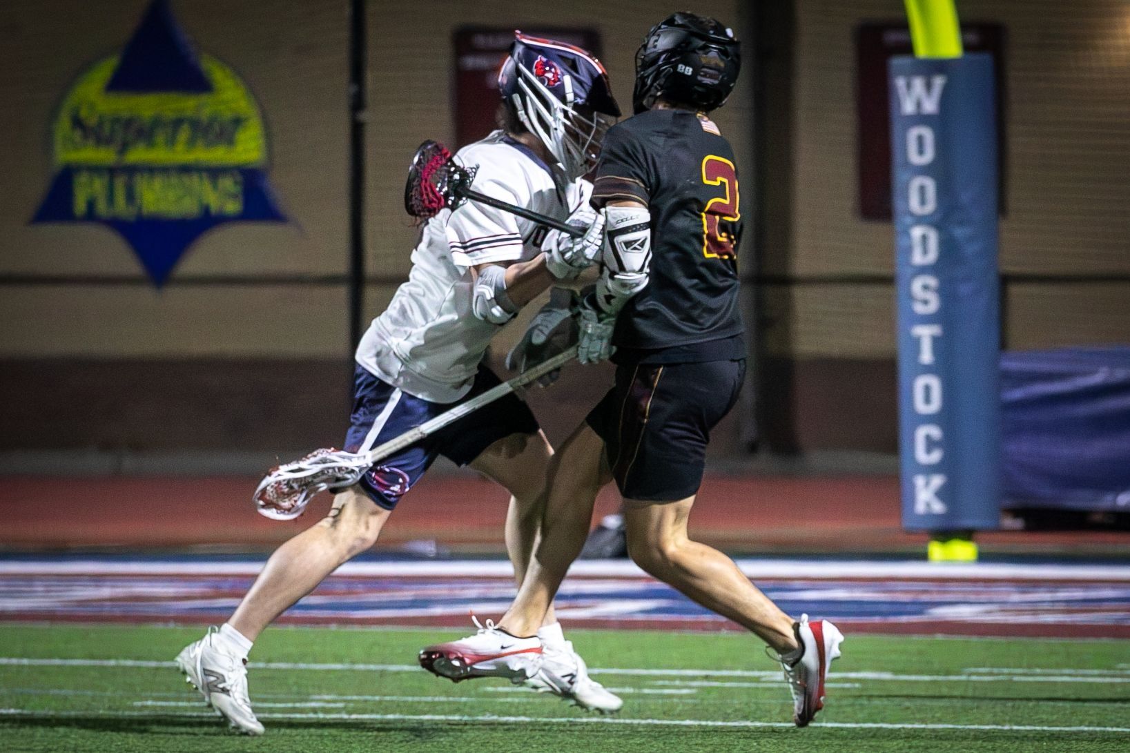 Two lacrosse players clash near the goal, one in white and one in black, on an indoor turf field.
