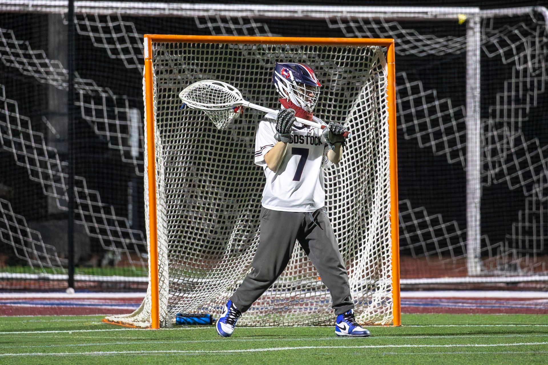 Lacrosse goalie in white jersey and helmet blocking the net on a field at night