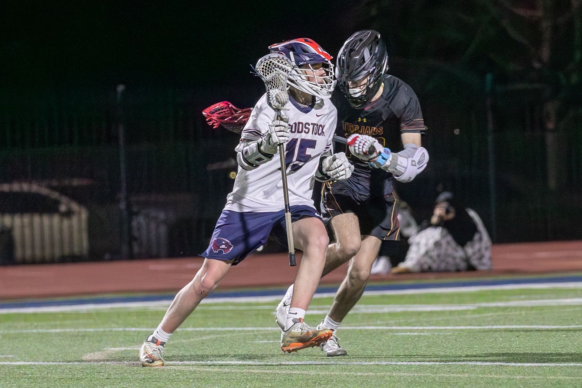 Two lacrosse players in white and black uniforms battling for the ball on a lit field at night