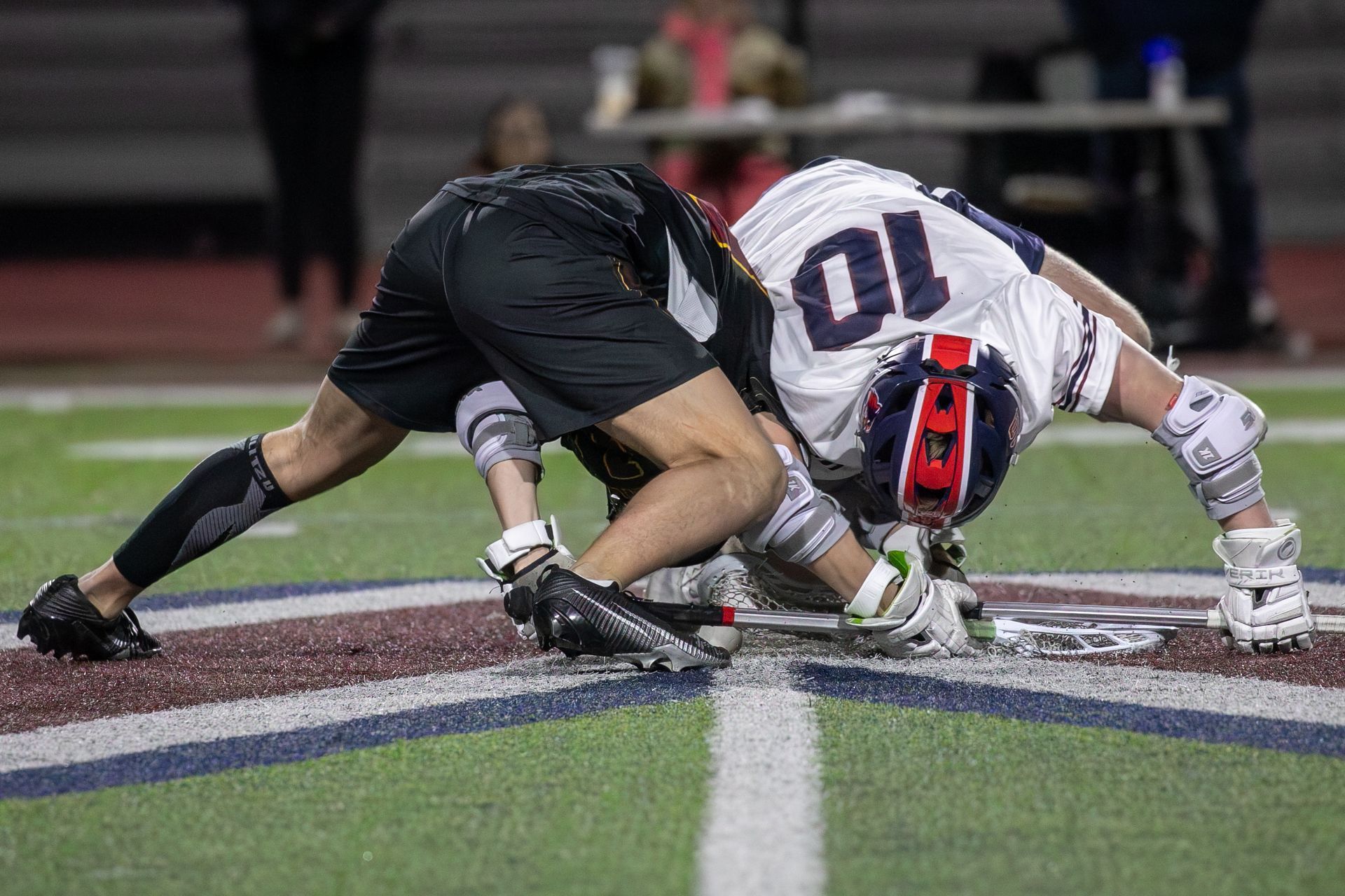 Two lacrosse players in black and white jerseys battle for the ball on a field at night.