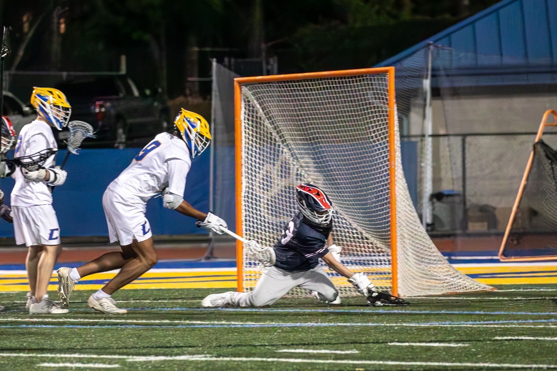 Lacrosse goalie blocking a shot at the goal while two players in white watch nearby at night.