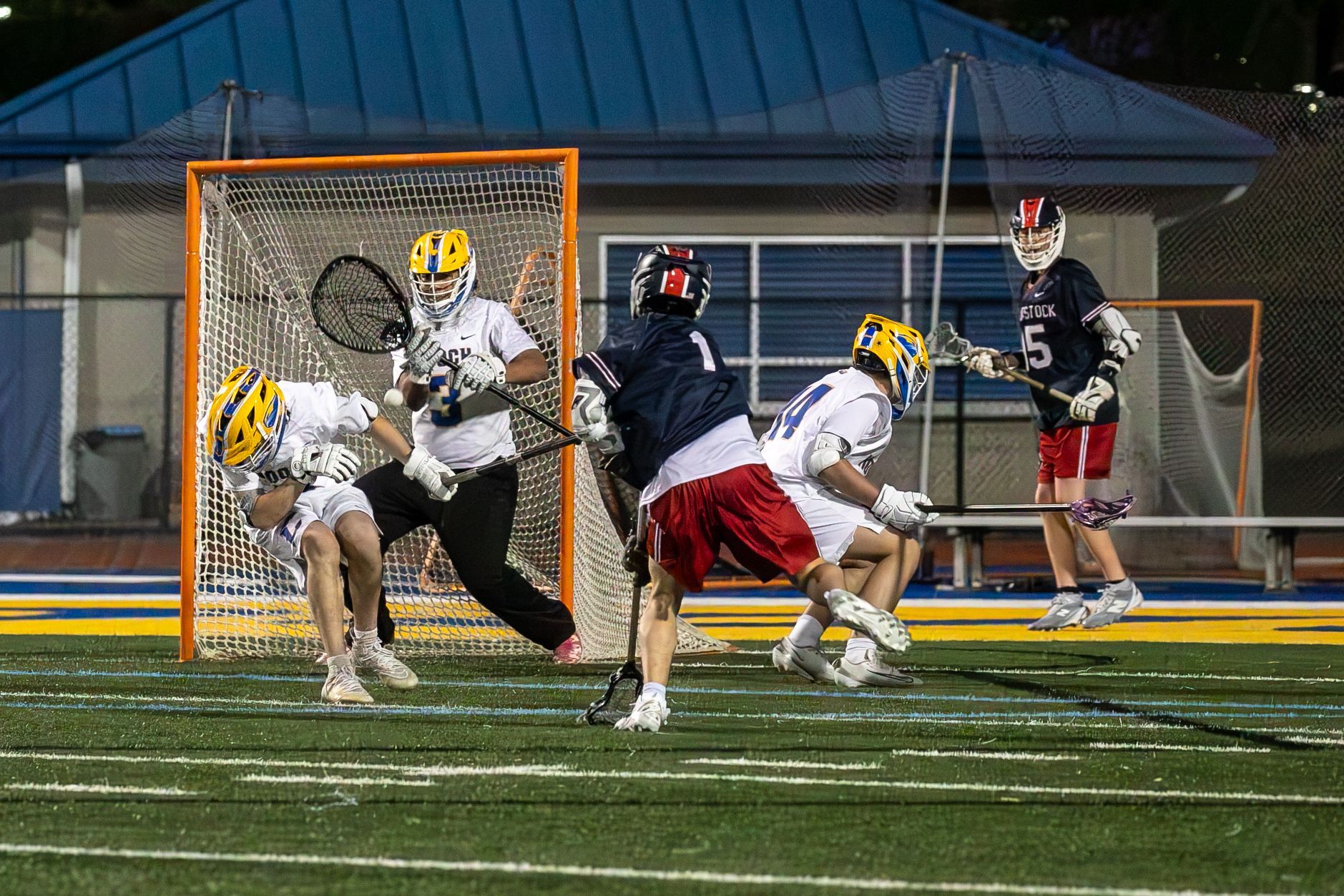 Lacrosse players in white and dark uniforms clash near the goal on a lit field at night