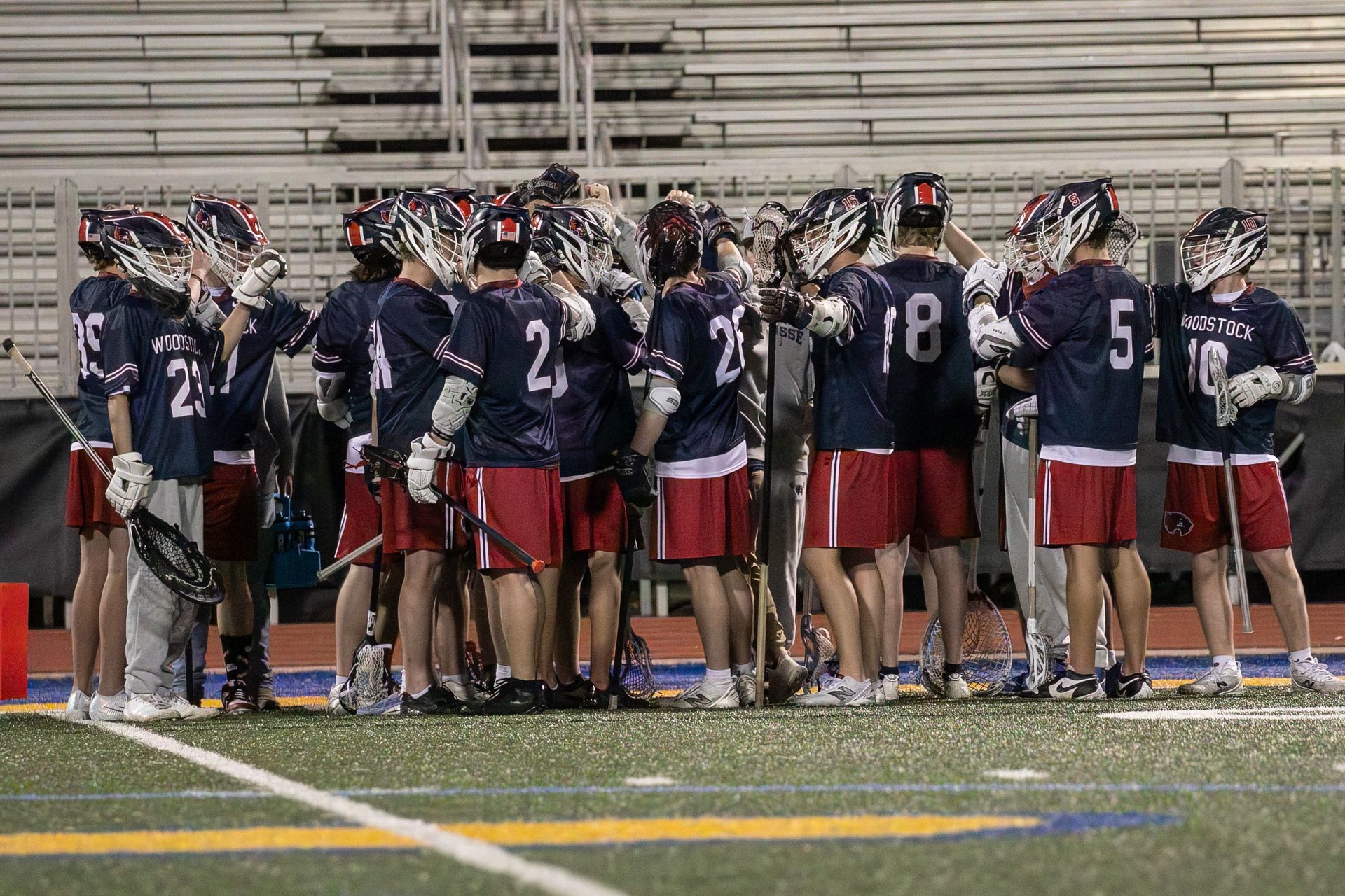 Lacrosse team huddled on the sideline at night, wearing navy jerseys and red shorts.