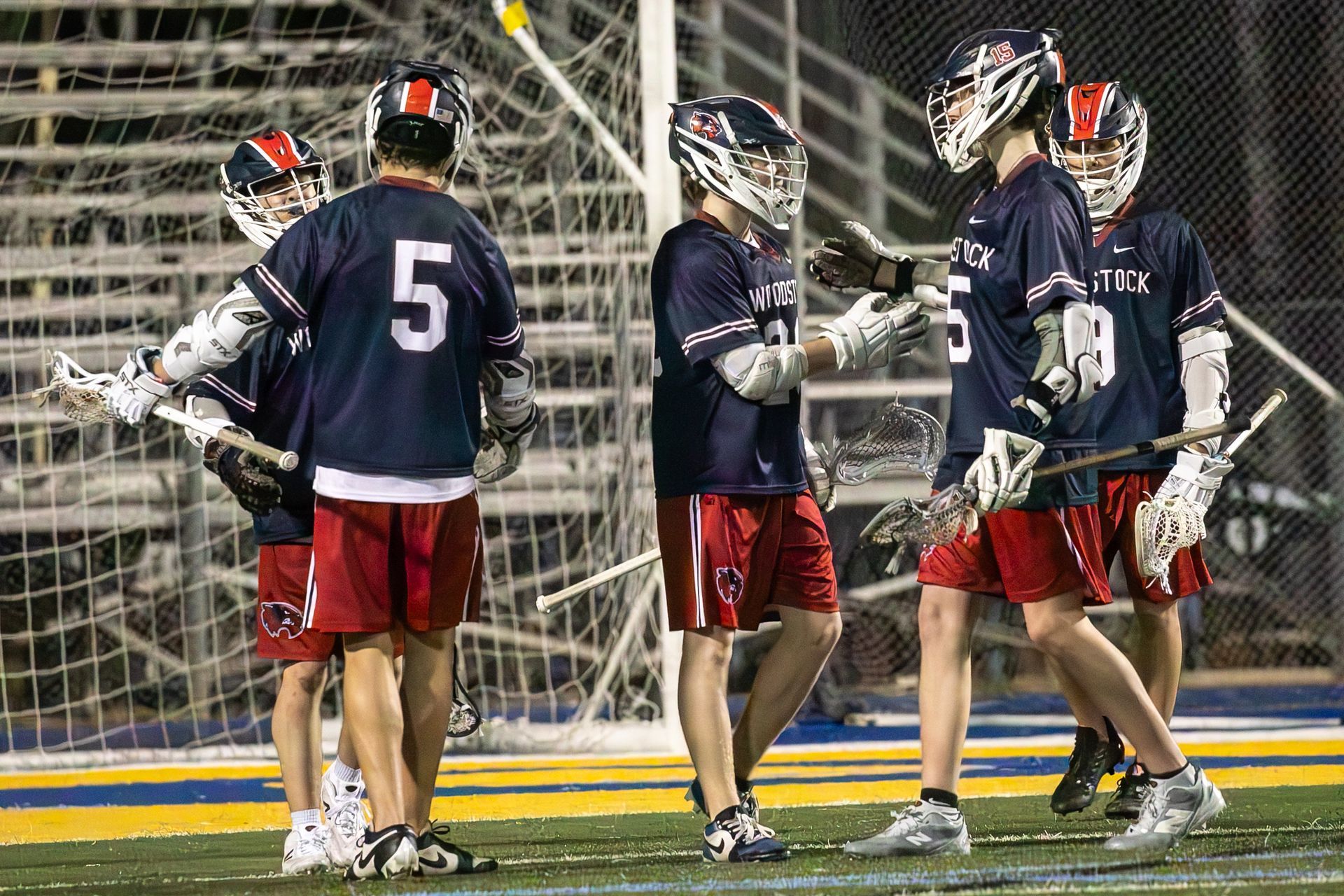 Lacrosse players in navy and red uniforms huddle near the goal on a lit field.