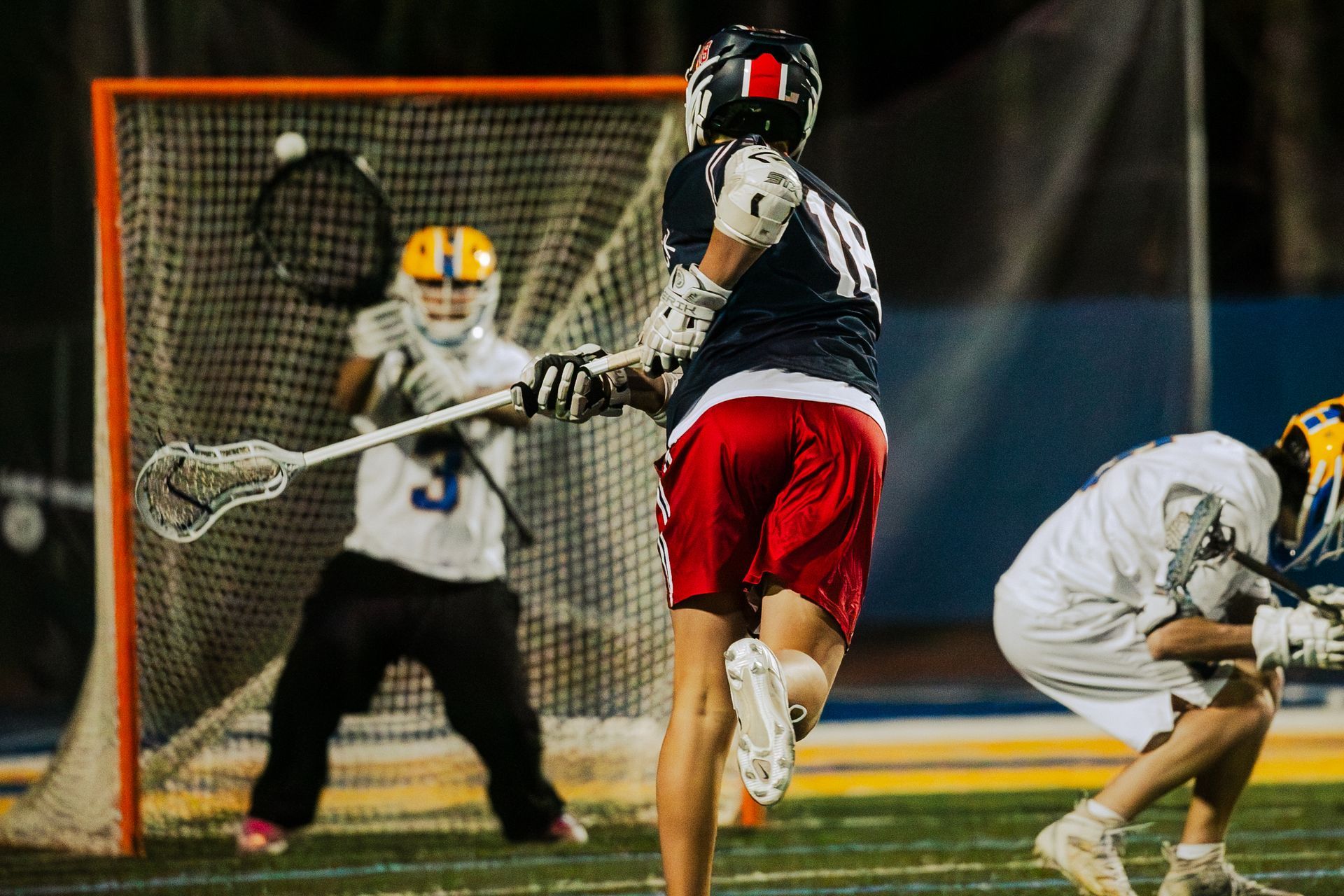 Lacrosse players in action near the goal, with a goalie in front of the net at night.