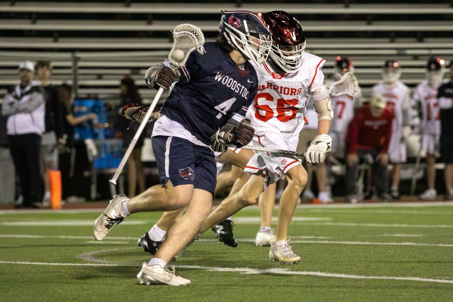 Two lacrosse players sprinting on a field, one in dark navy and one in white and red, at night
