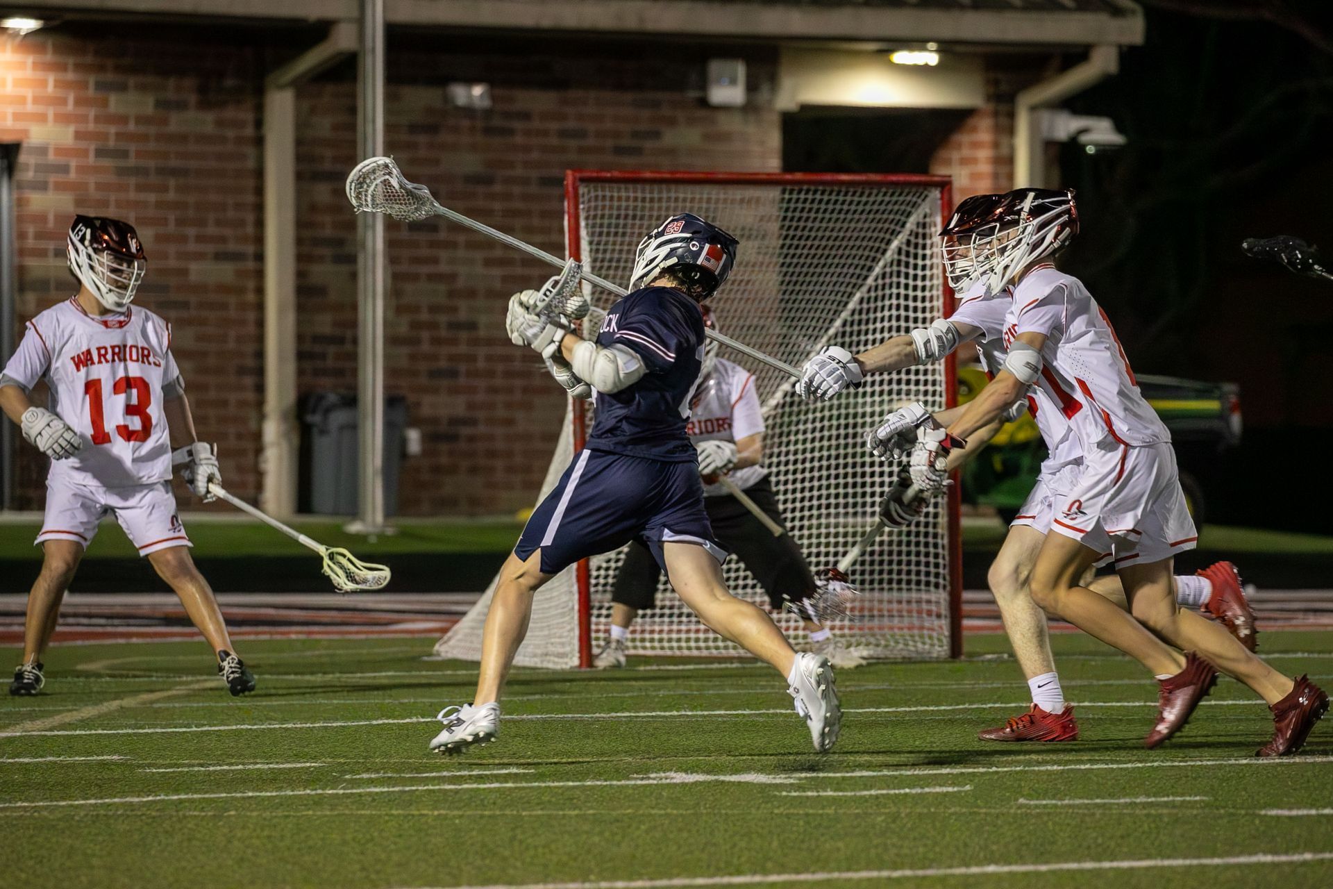 Lacrosse players in navy and white jerseys battle for the ball near the goal at night.