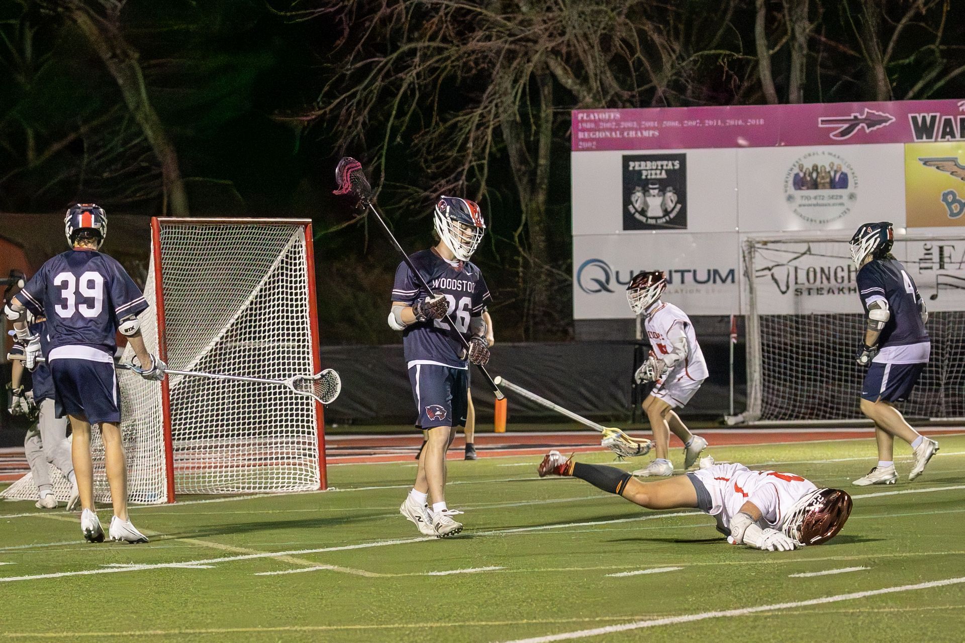 Lacrosse game at night near the goal; players cluster as one lies on the turf.