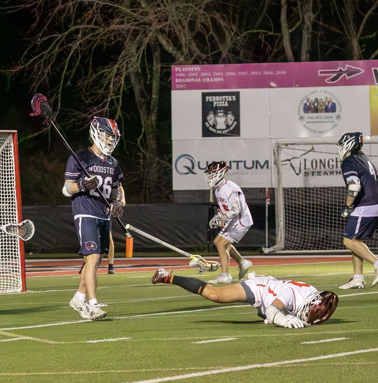 Lacrosse players near the goal, with one player lying on the field after a play at night.