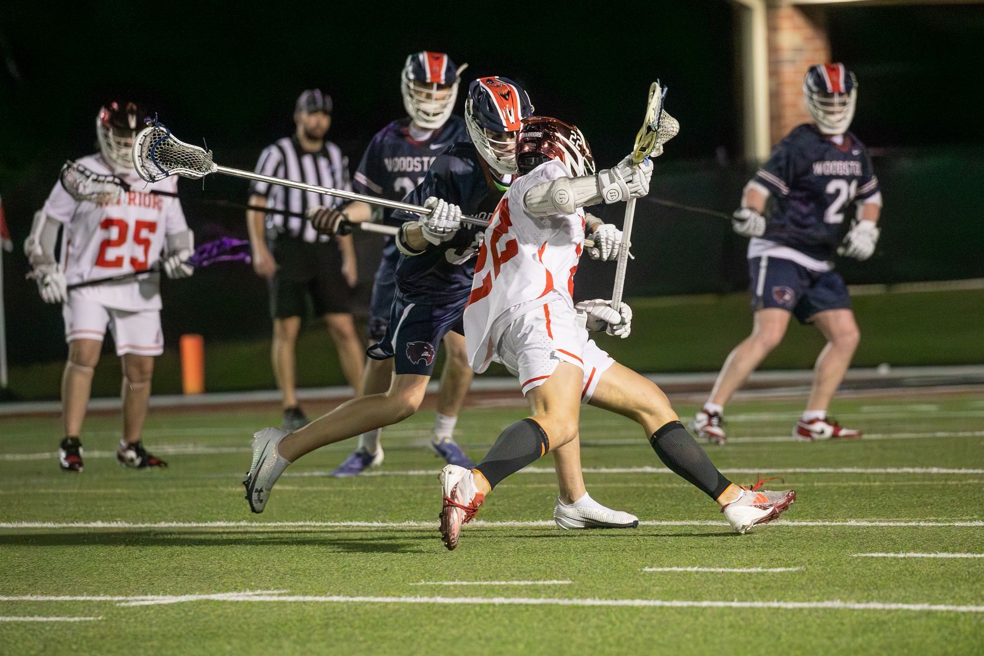 Lacrosse players in white and navy compete for the ball under stadium lights at night.