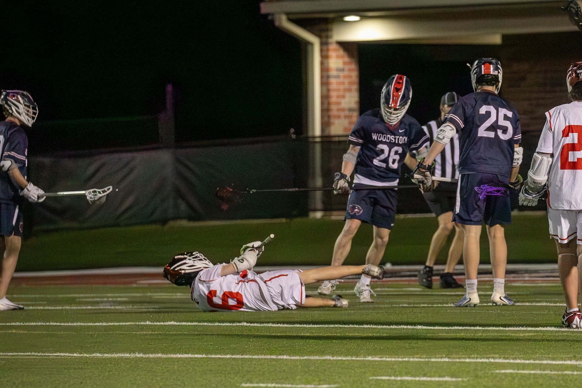 Lacrosse player in white lies on turf as navy-clad players stand nearby under stadium lights