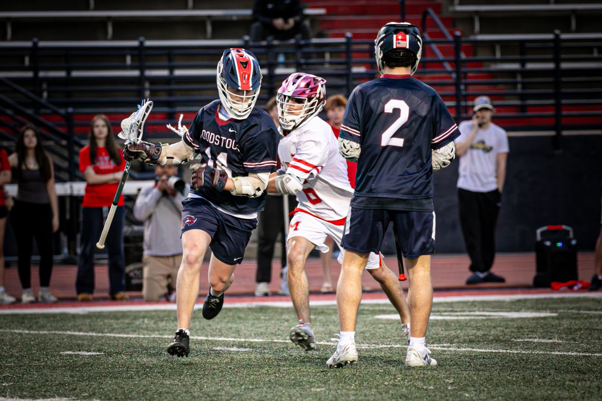 Three lacrosse players in dark and white uniforms compete on an indoor field