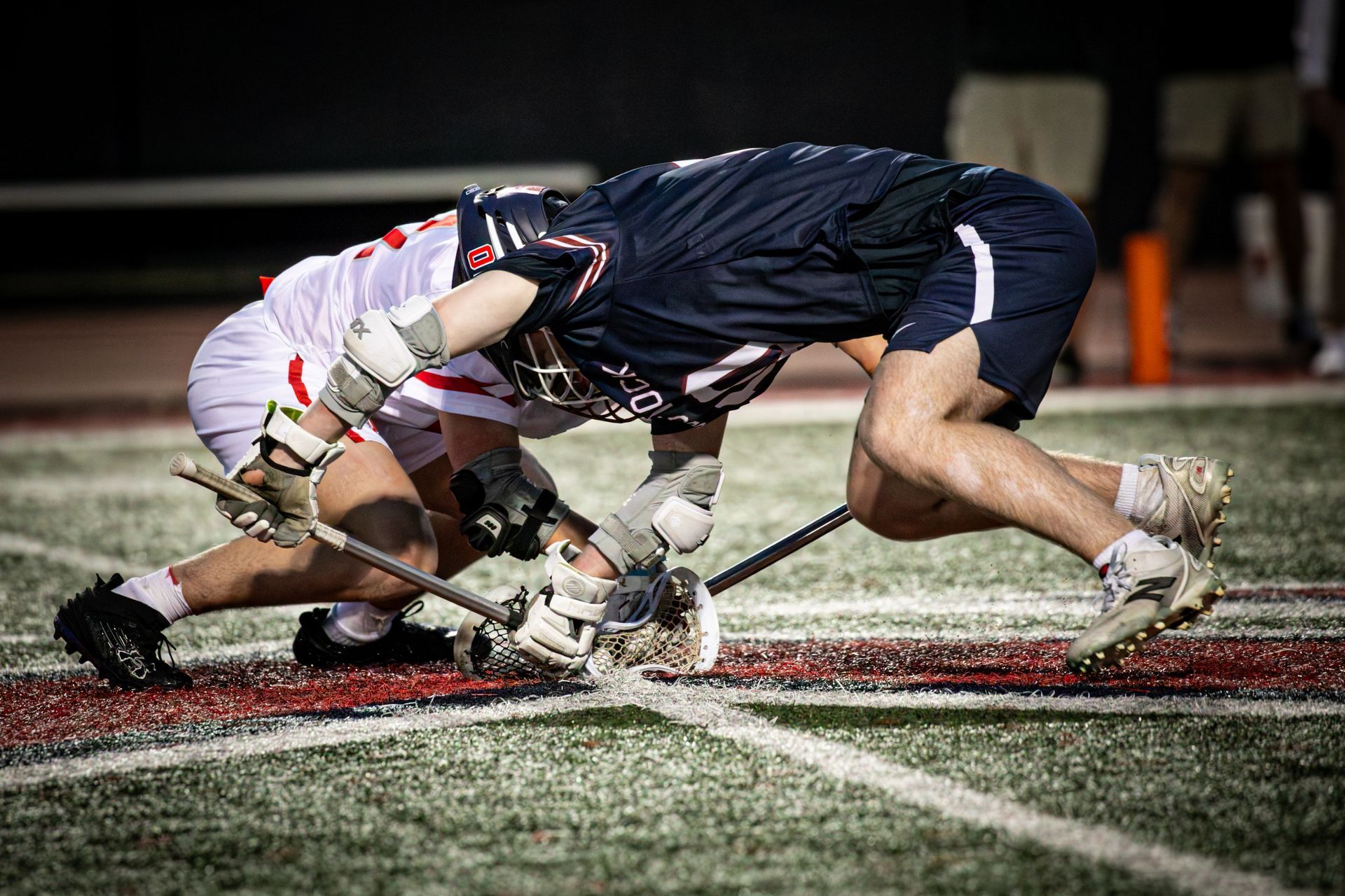 Two lacrosse players collide while battling for the ball on a turf field at night.