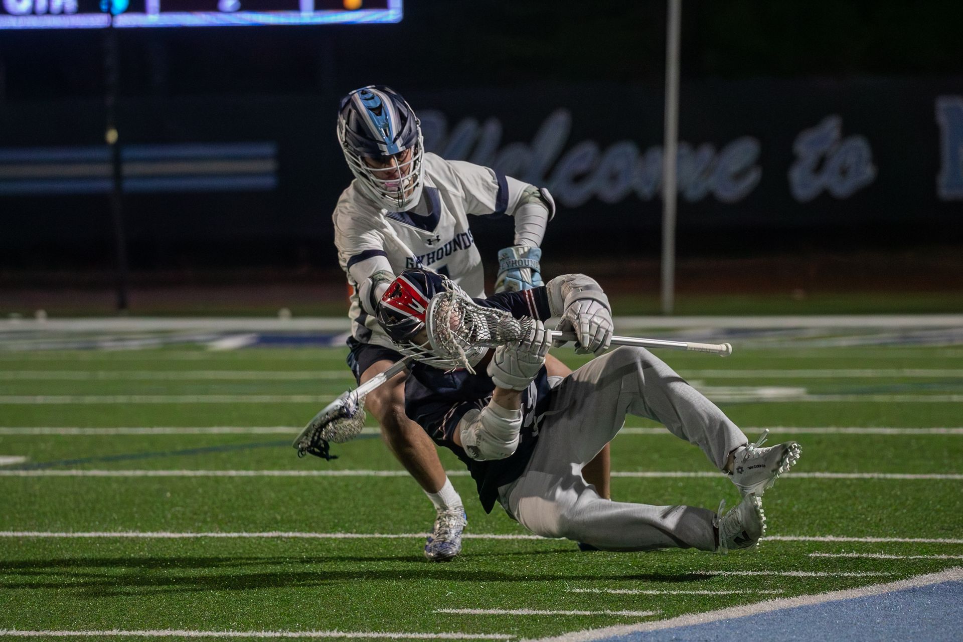 Two lacrosse players collide on a lit field, one tackling the other near the sideline at night