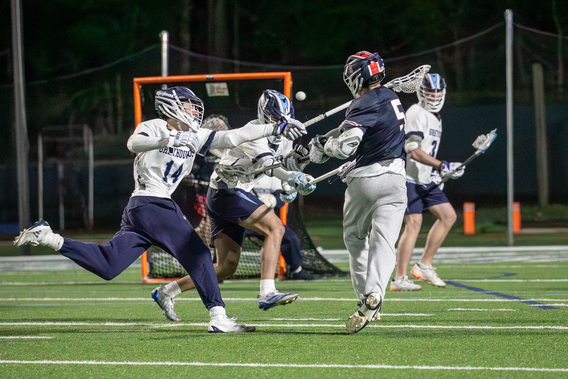 Lacrosse players in navy and white compete on a lit field at night, one leaping to catch the ball.