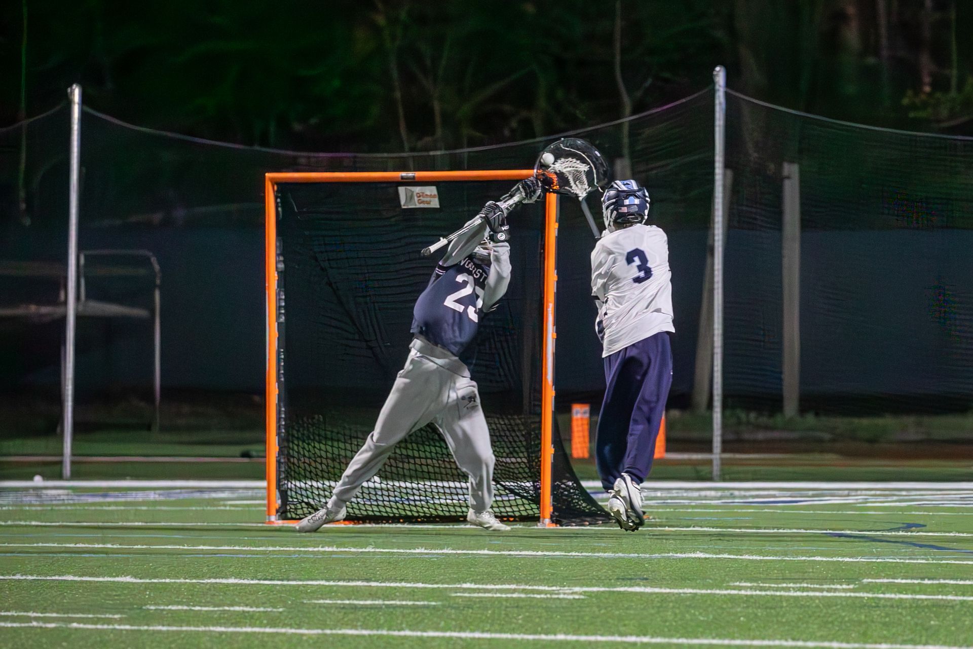 Two lacrosse players contest a shot near the goal at night on a lit field