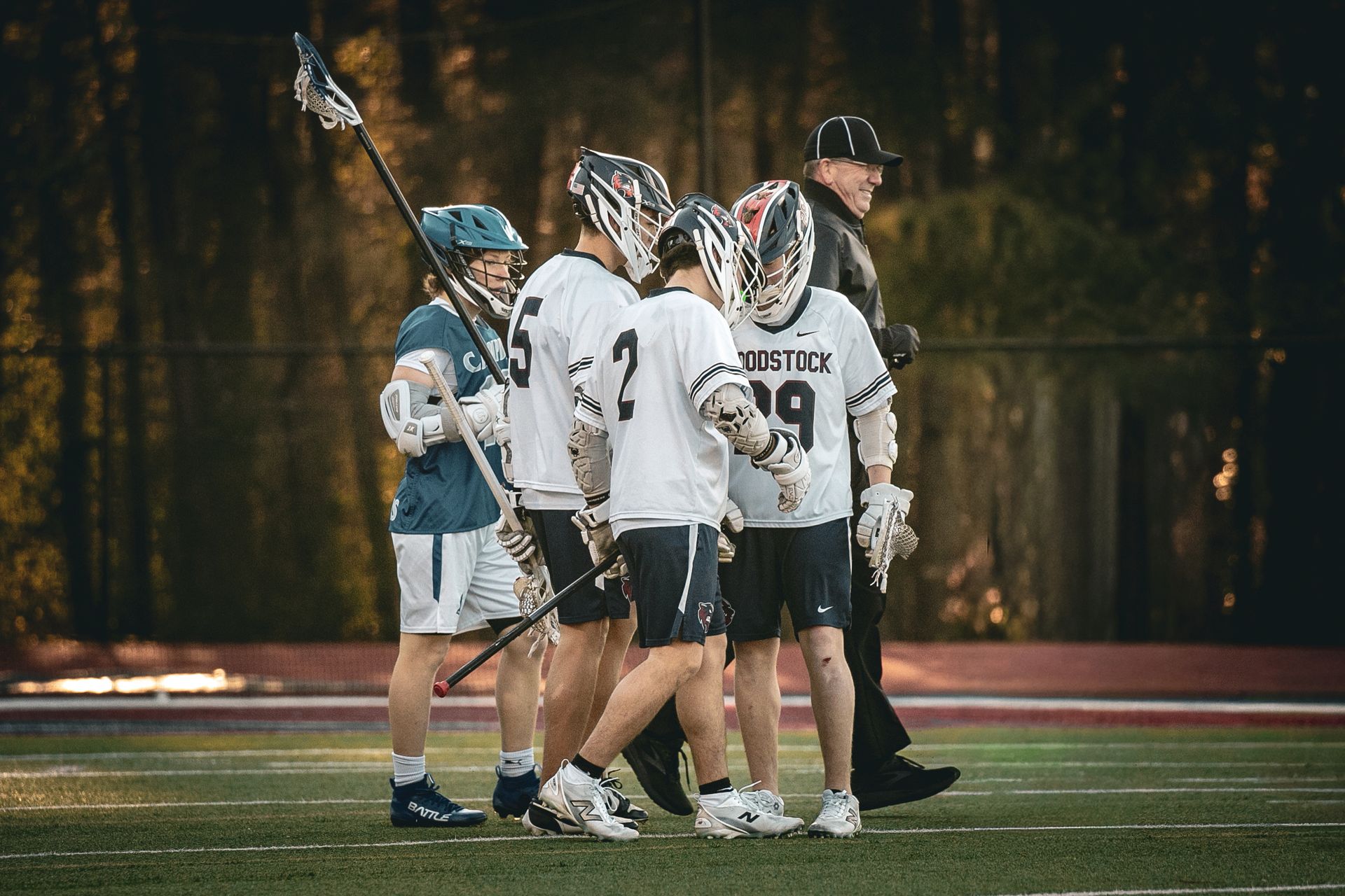 Lacrosse players huddle on a field at night with a coach beside them.