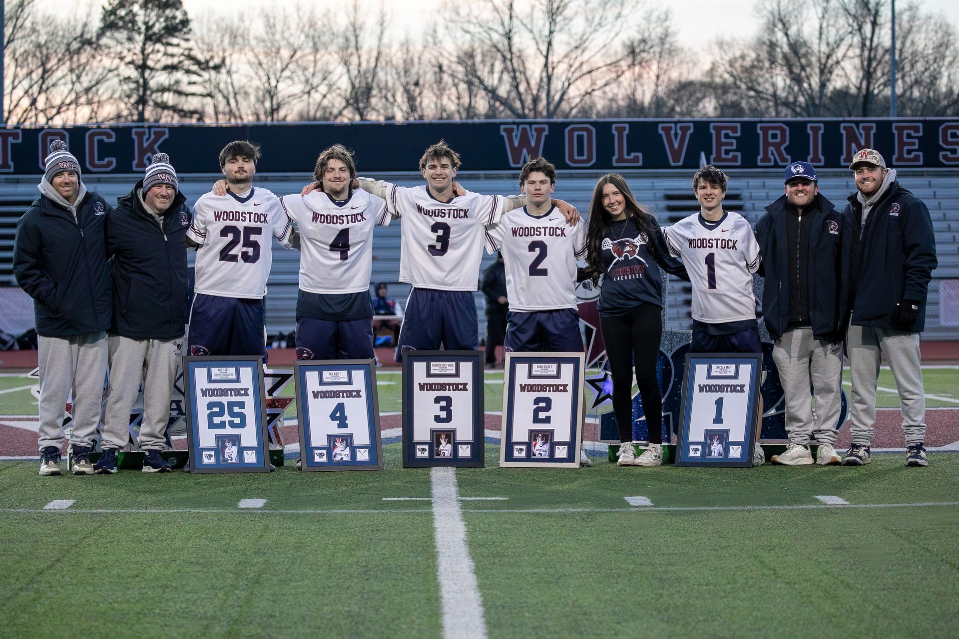 Wolverines football players and coaches stand with senior night signs on a field at dusk.