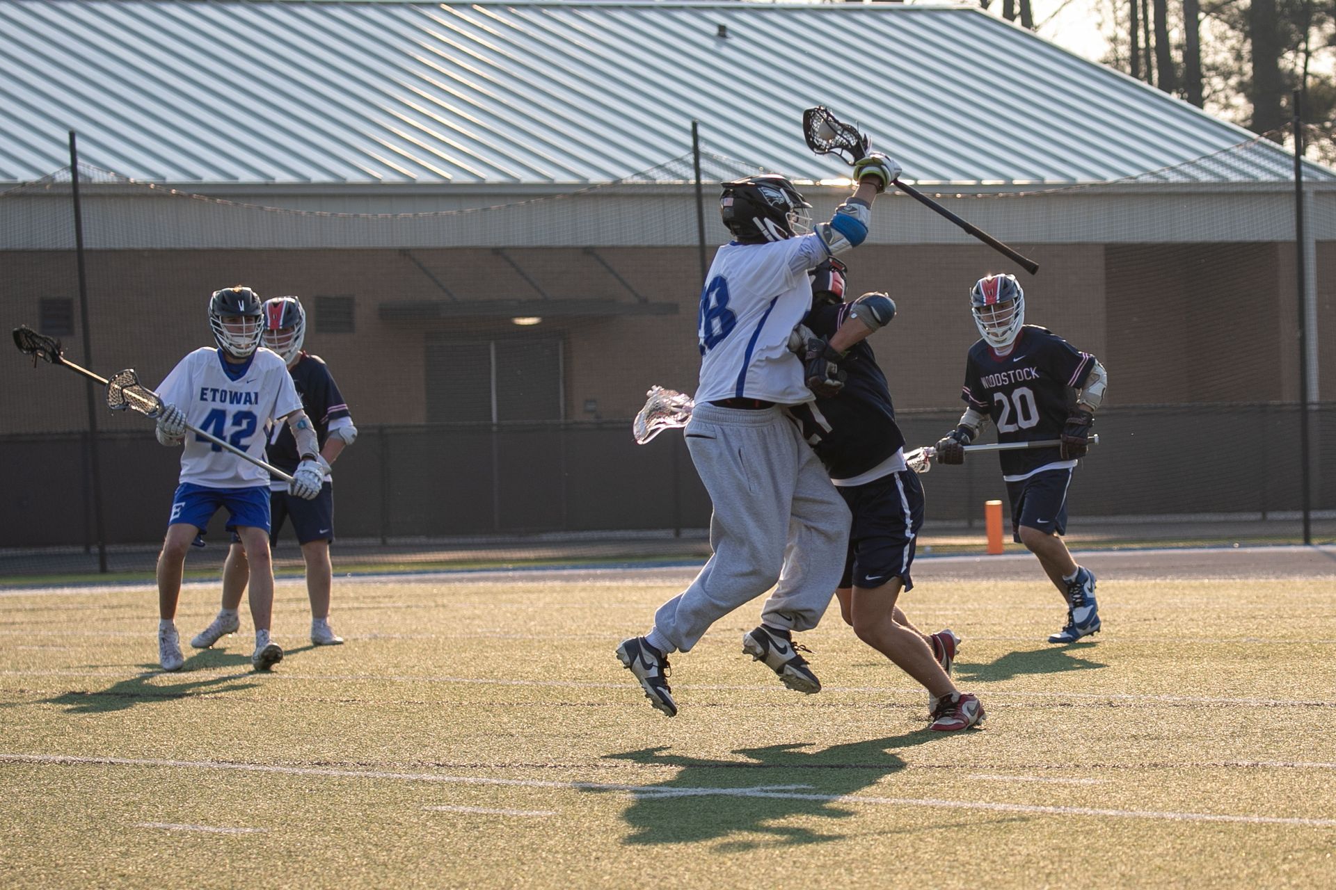 Lacrosse players in white and black jerseys leap for a contested ball on a sunny field.