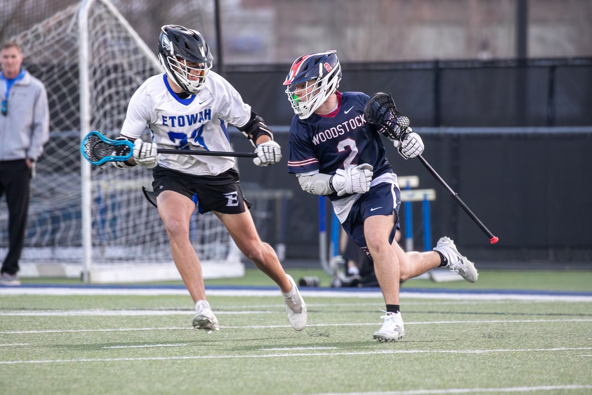 Two lacrosse players sprinting on a field, one in a white jersey and one in a dark jersey