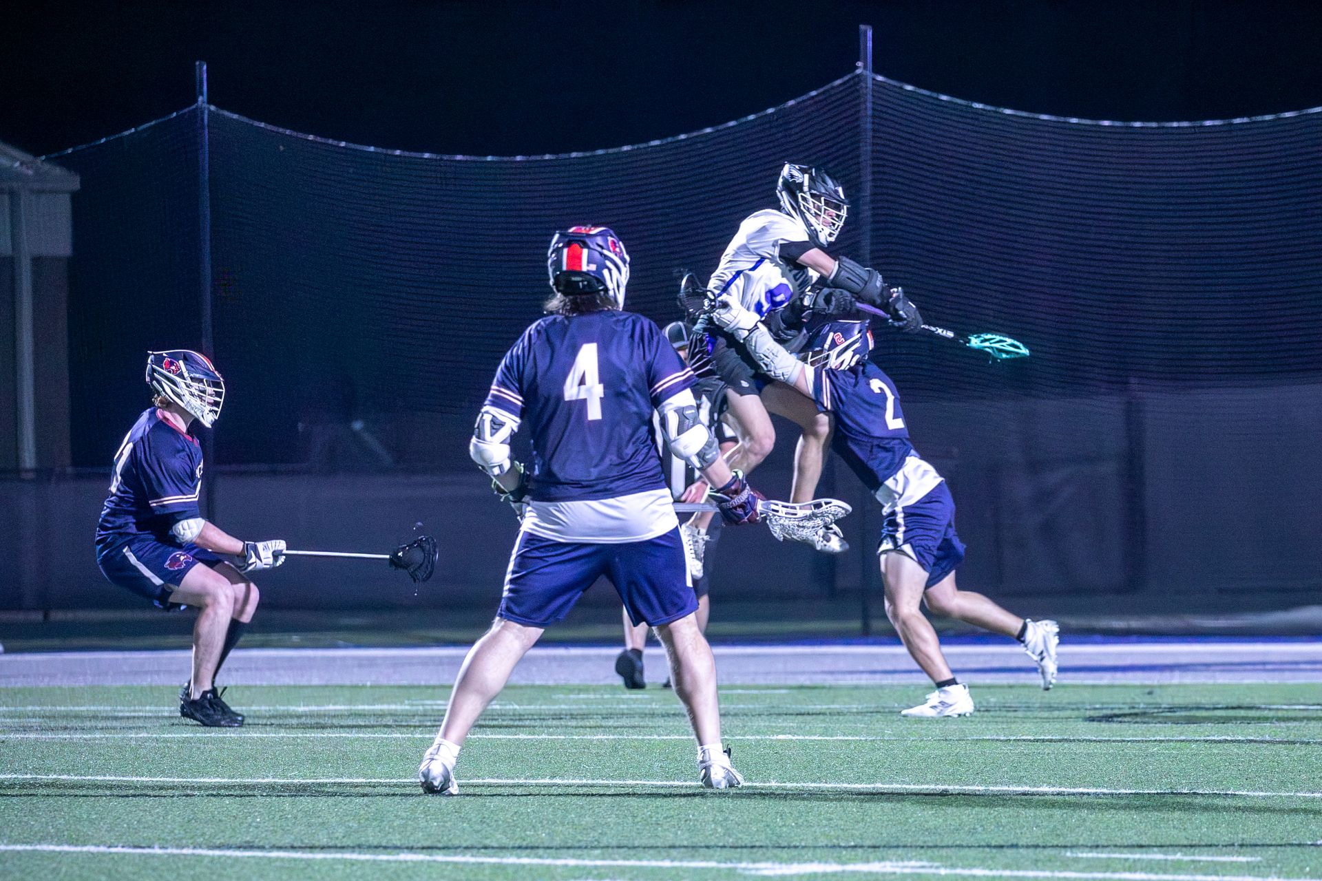 Lacrosse players in navy uniforms contest a high pass on a lit field at night.