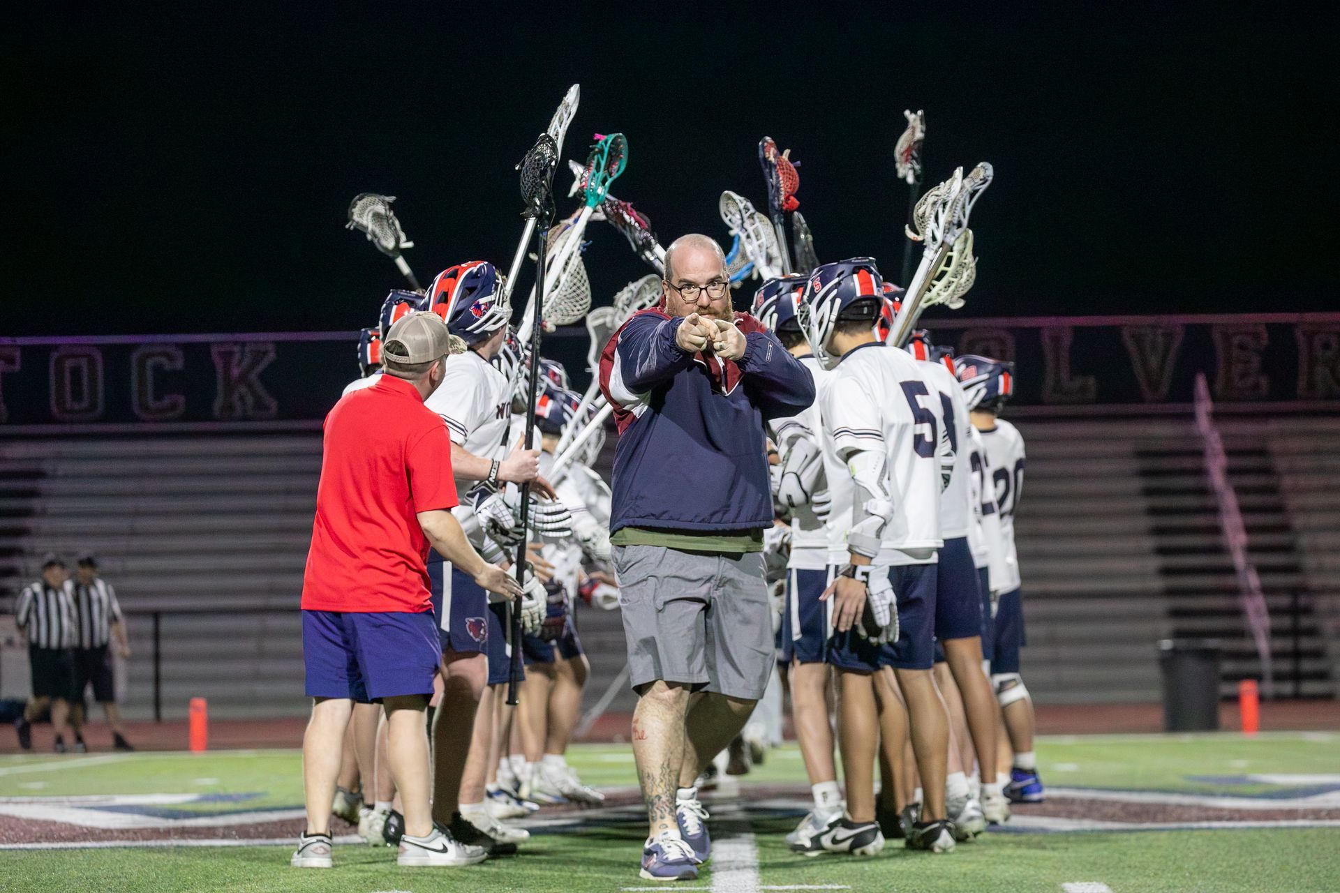 Lacrosse players huddle on a field at night while a man in a navy shirt walks toward the camera.