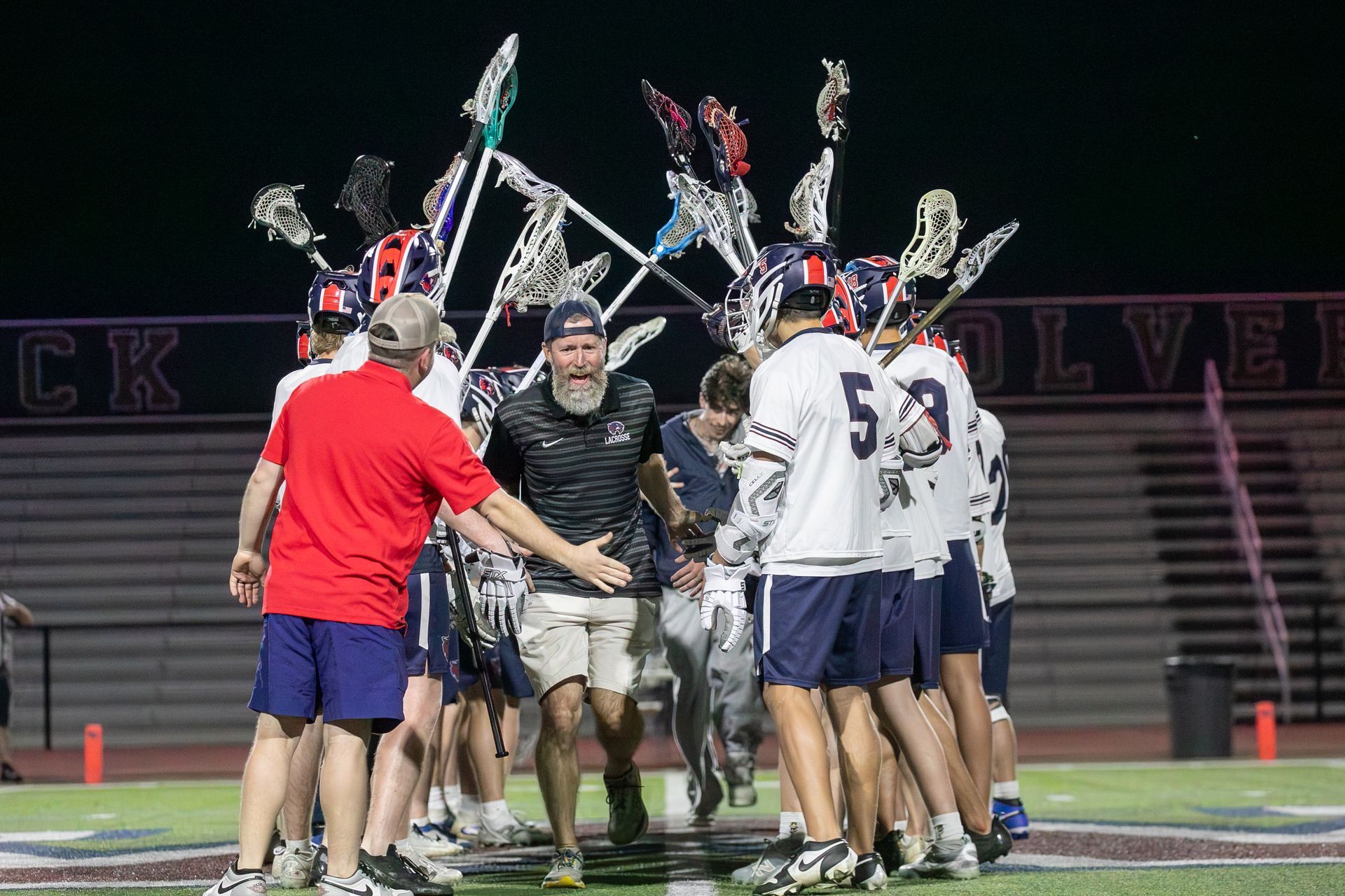 Lacrosse players huddle on a lit field after a night game, with a coach in red joining the celebration.