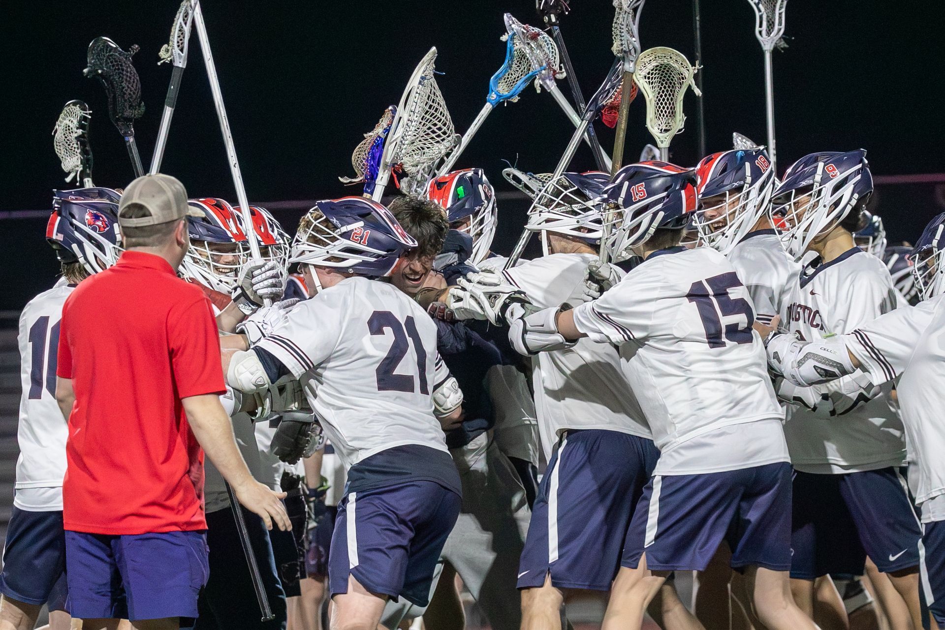 Lacrosse team in white jerseys huddles under stadium lights, raising sticks before a game