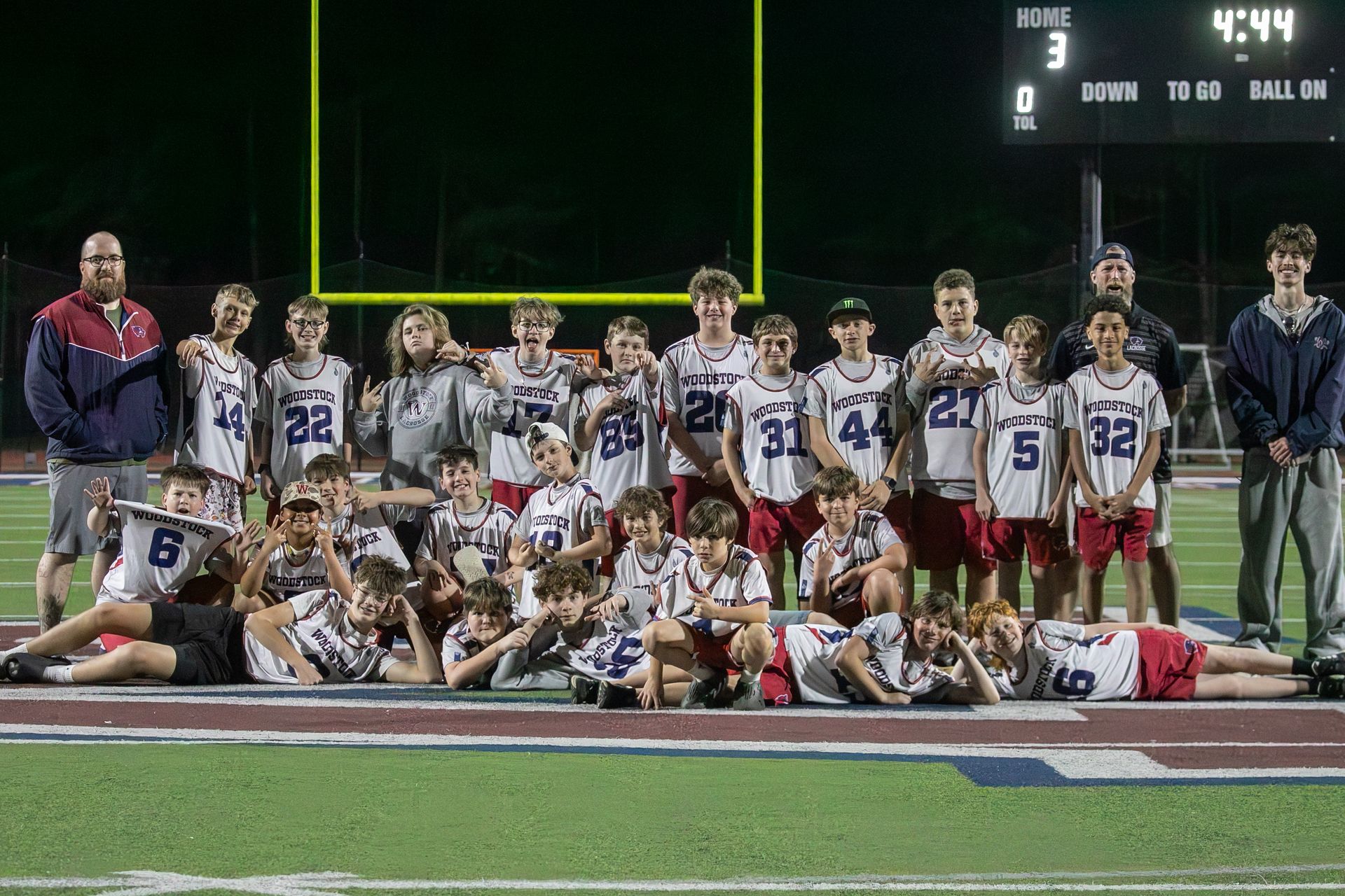 Youth football team posing on a field at night after a game, with goalposts and scoreboard in the background