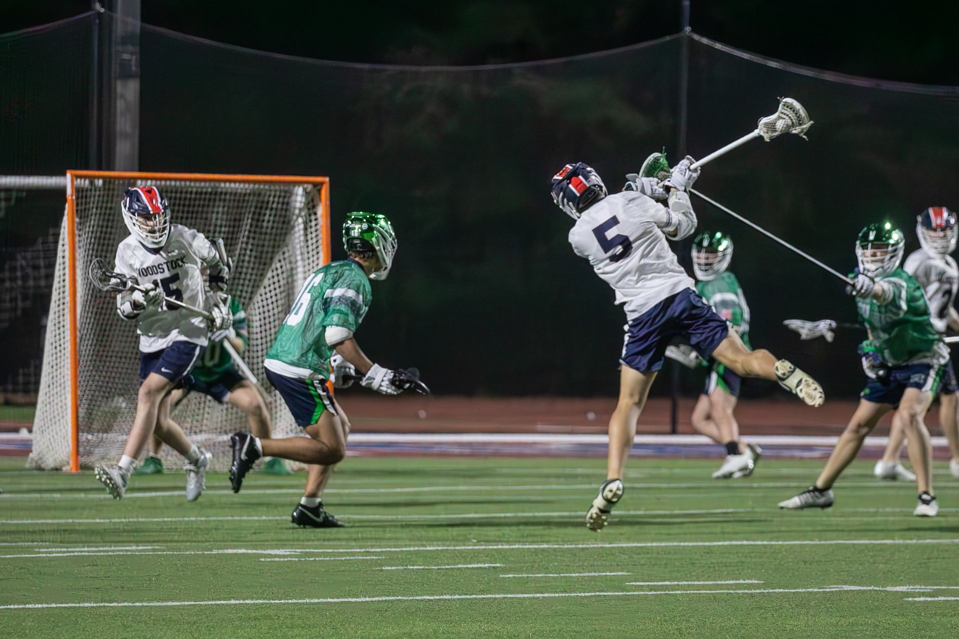Lacrosse game near goal, white attacker shoots as green defenders close in under stadium lights