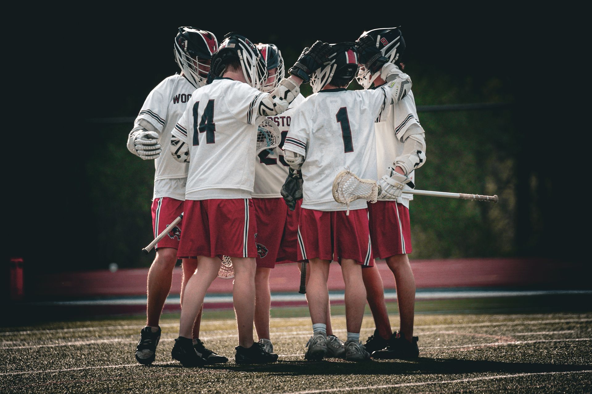 Lacrosse team huddled on a wet field at night, wearing white jerseys and red shorts.