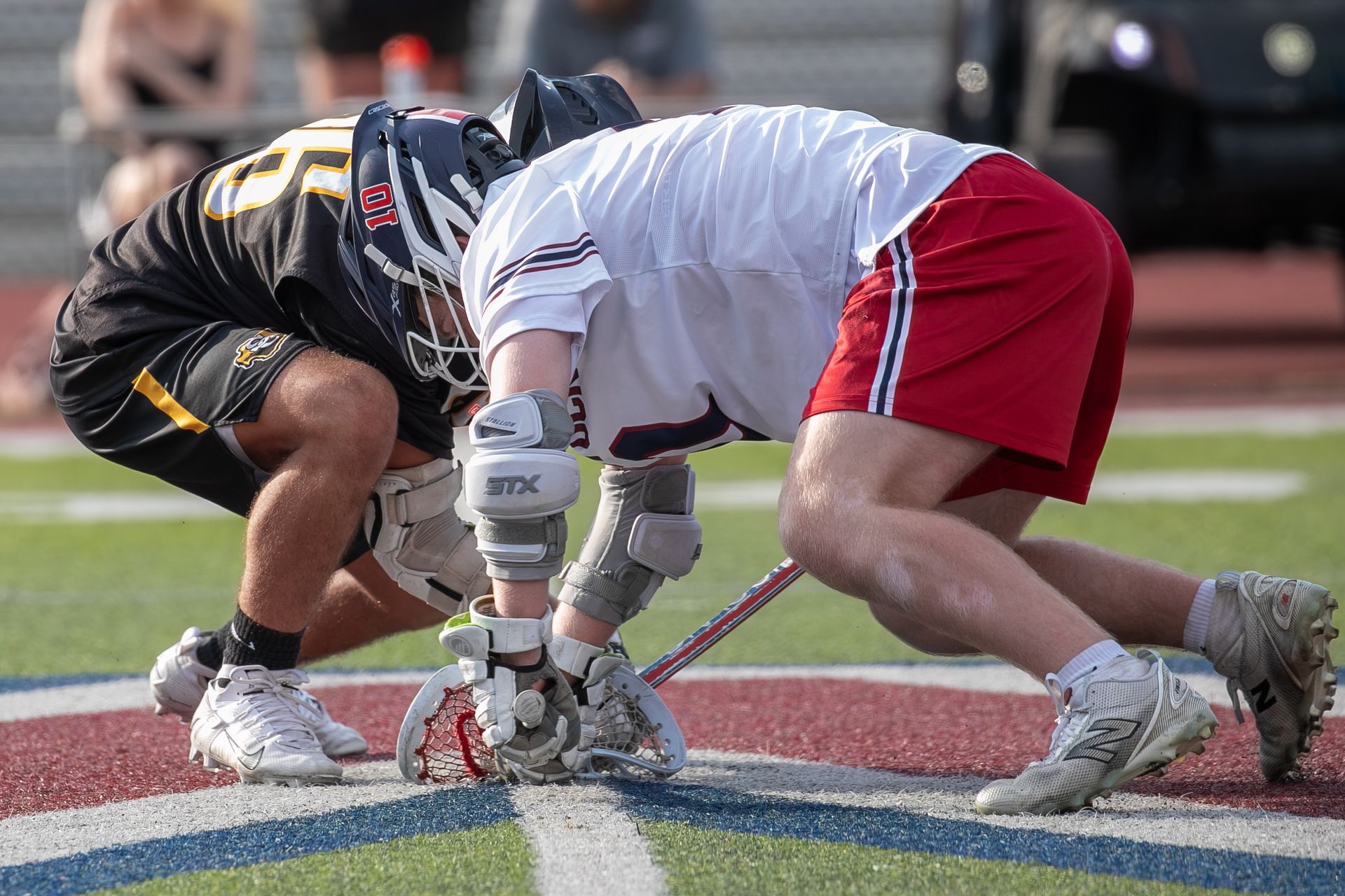 Two lacrosse players crouch at the faceoff line on a field, sticks pressed together.