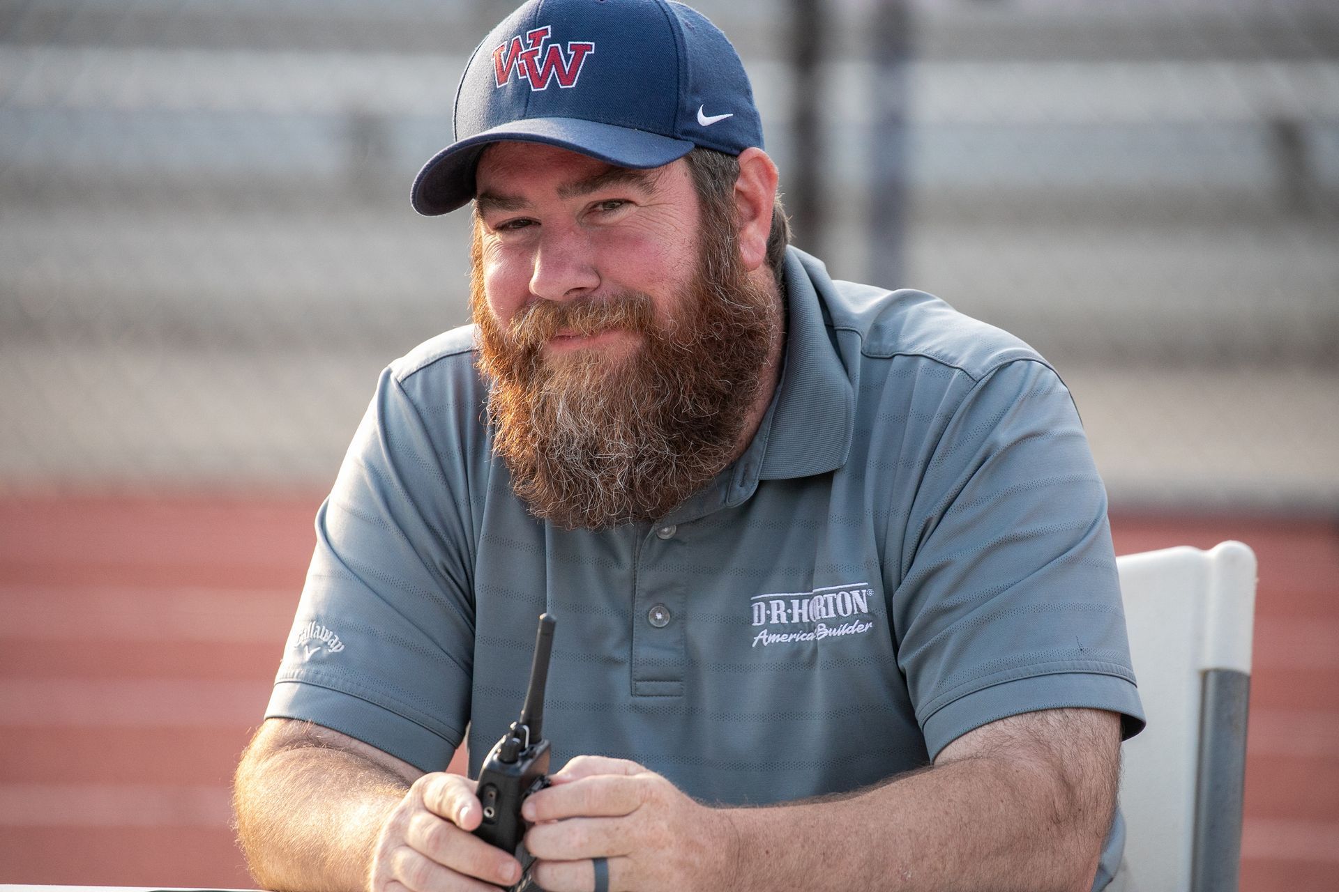 Bearded man in a blue cap and gray polo holding a radio outdoors, smiling at a sports venue