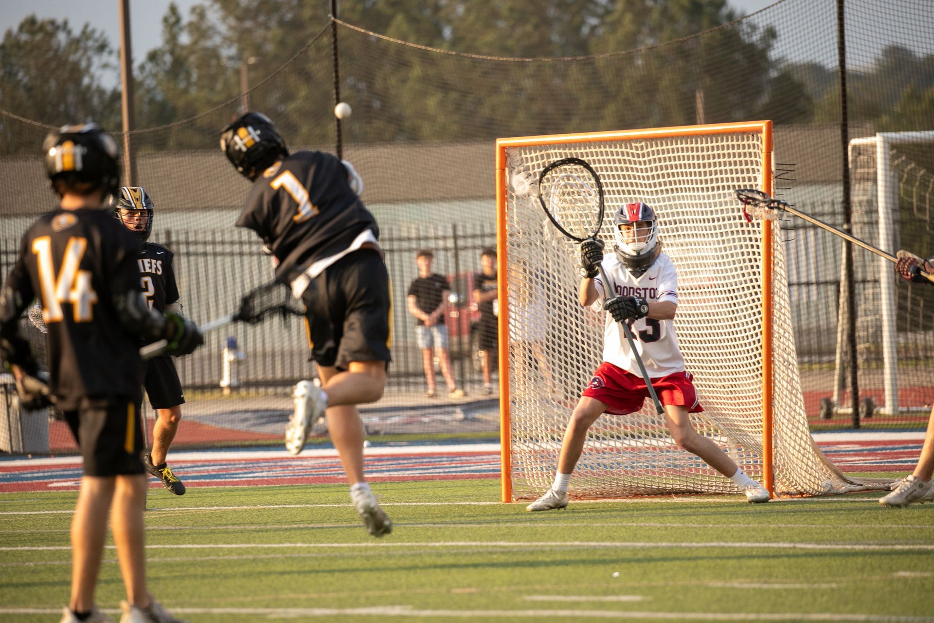 Lacrosse player in white and red defending the goal as two black-uniformed players approach on a field