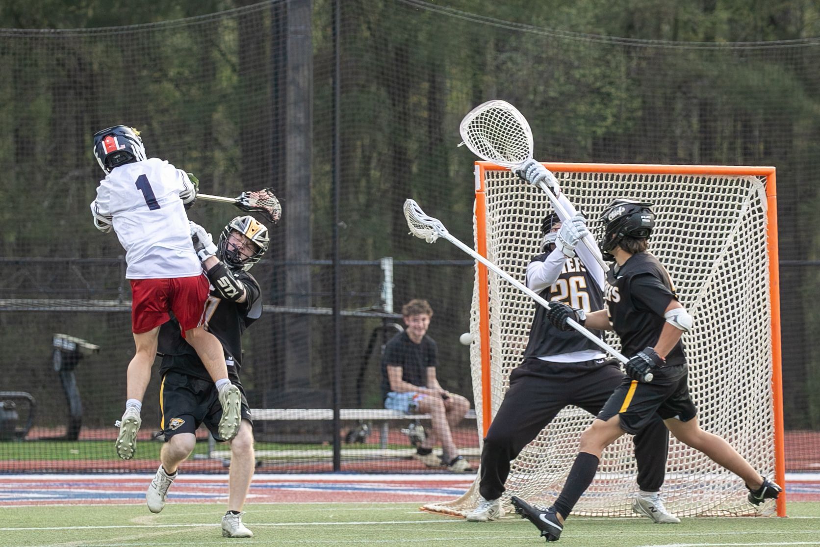 Lacrosse players contest a shot near the goal on an outdoor field.