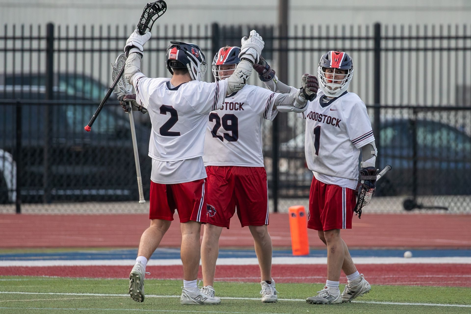 Three lacrosse players in white and red uniforms celebrate on the field with sticks raised.